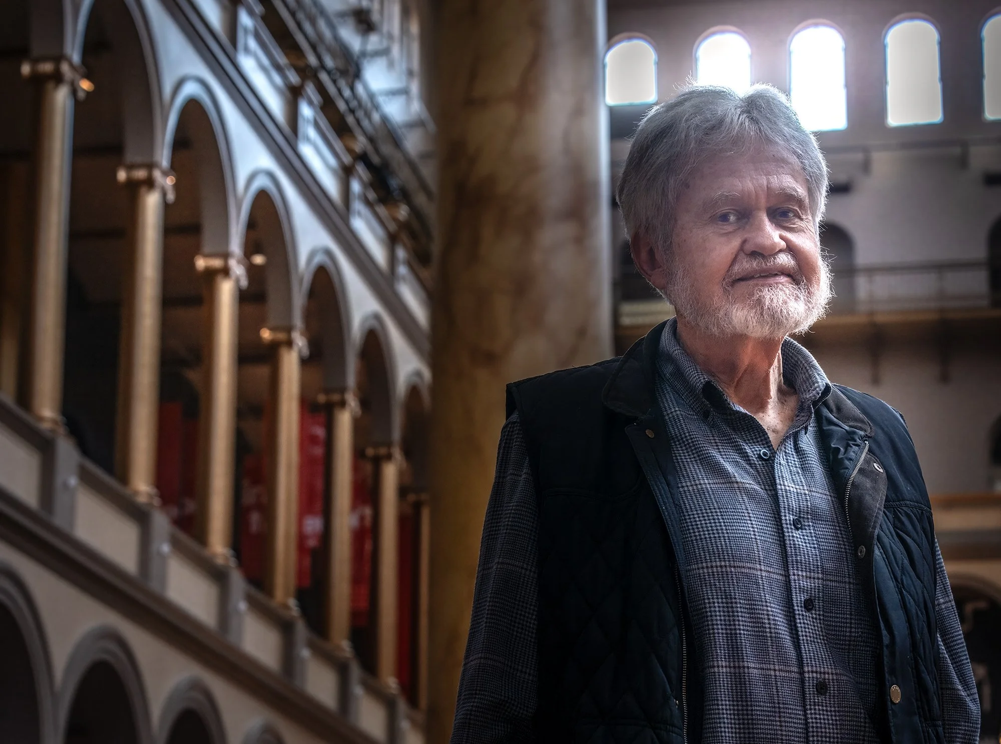 An older man with gray hair and a beard standing in a building with arched windows and bookshelves, looking toward the camera.