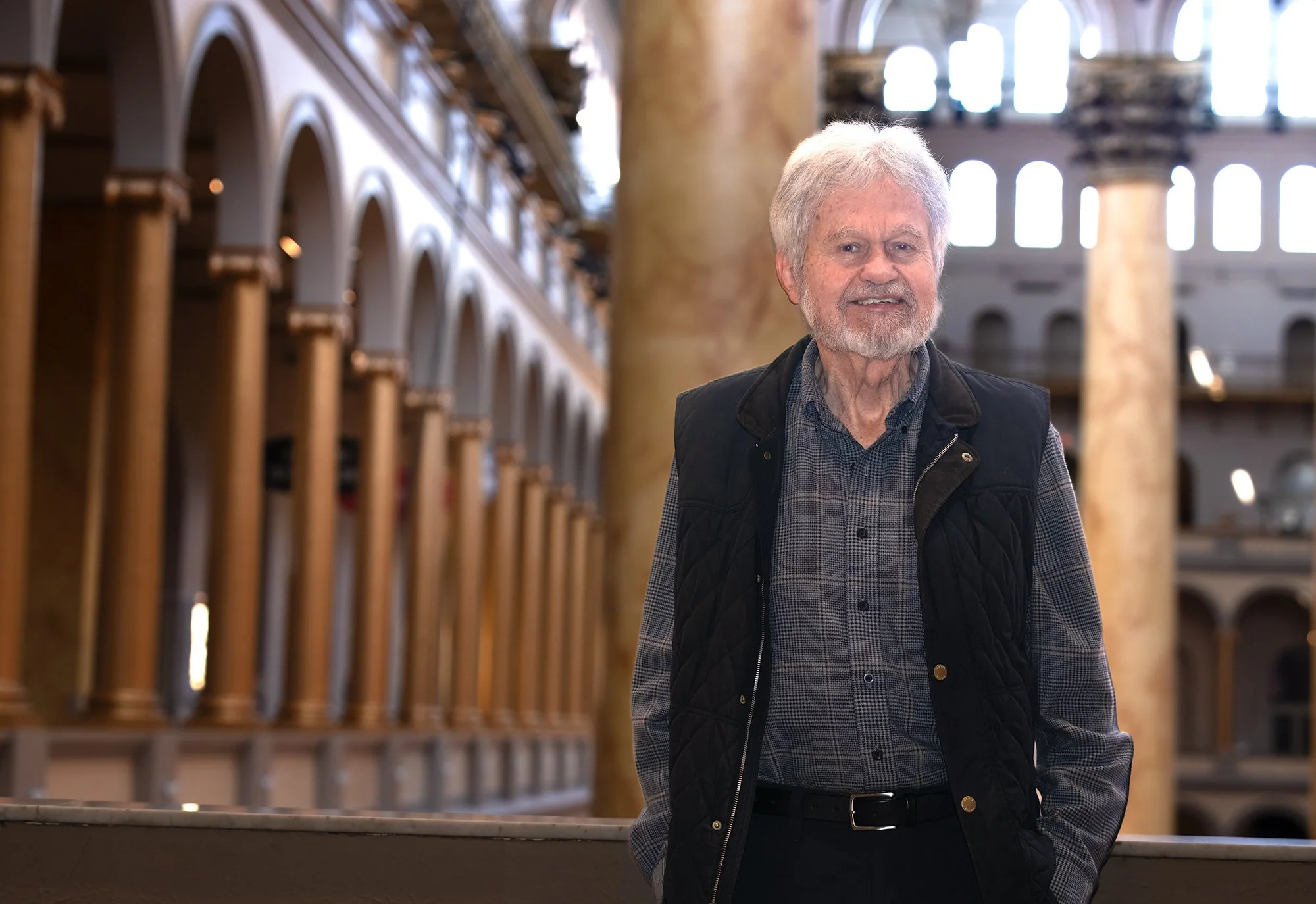 Older man with white hair and a beard standing inside a large building with tall windows and wooden shelves.