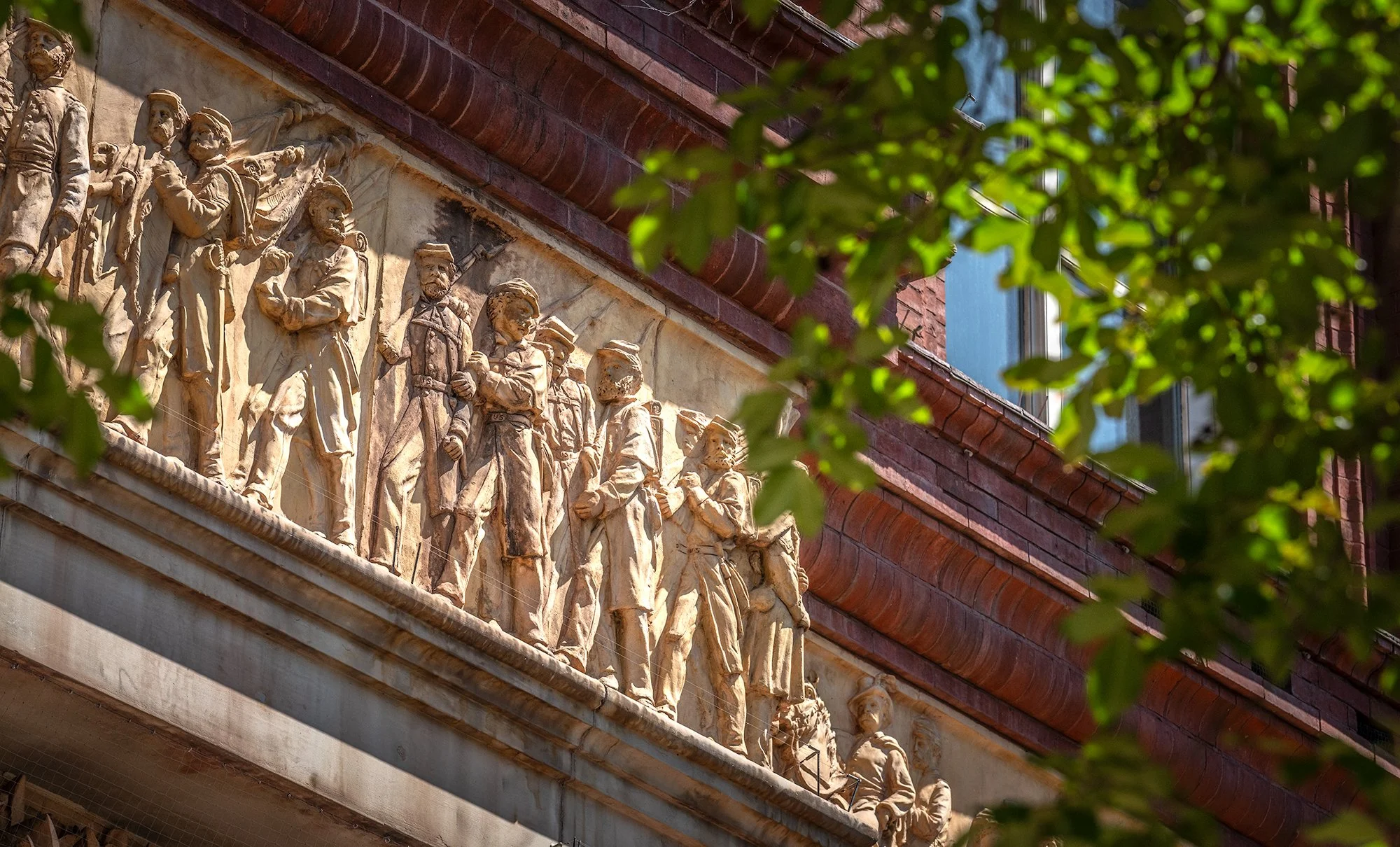 Stone relief sculpture of historical figures on a building, partially obscured by green leaves, with brick and window in background.