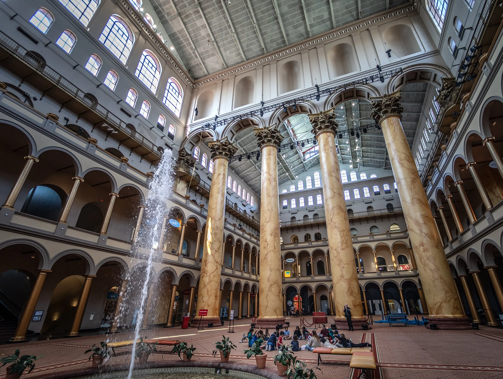 Interior of a grand historic building with large columns, multiple levels of arched windows, and a central fountain with water spray. People are sitting on the floor near the fountain, and some are walking around.