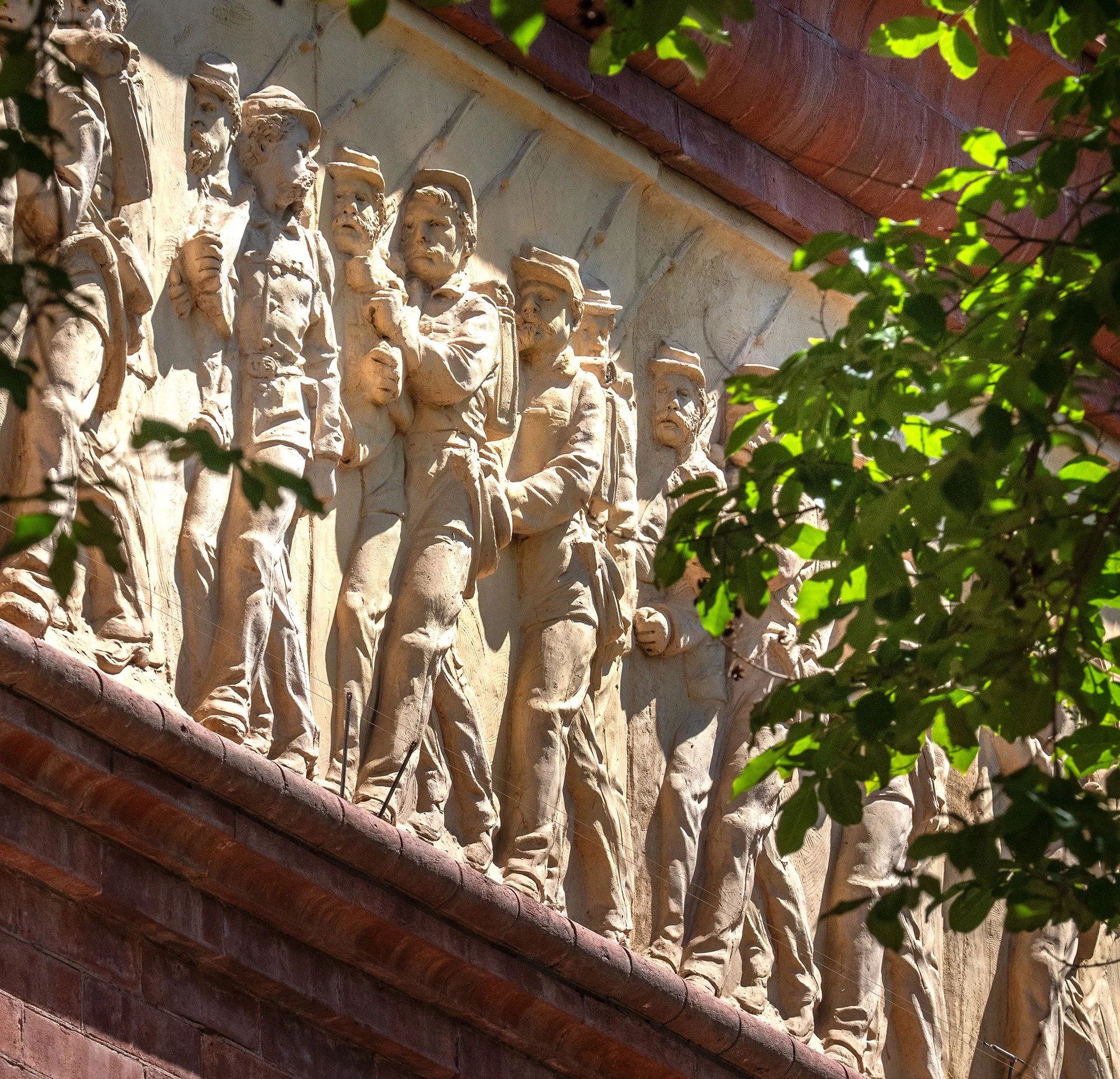 Close-up of beige stone sculptures depicting historical figures on a building's exterior wall, partially obscured by green foliage.