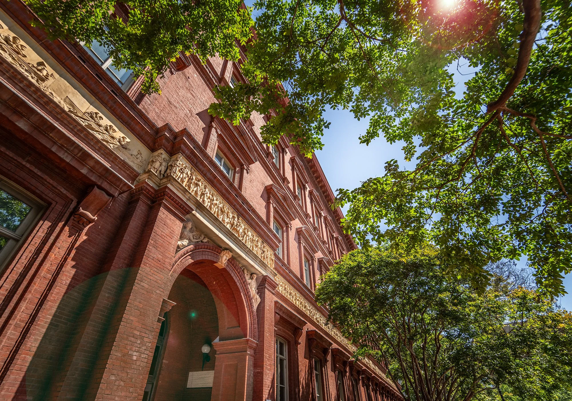 Red brick historic building with ornate architectural details, archways, and statues, surrounded by lush green trees under a clear blue sky with sunlight