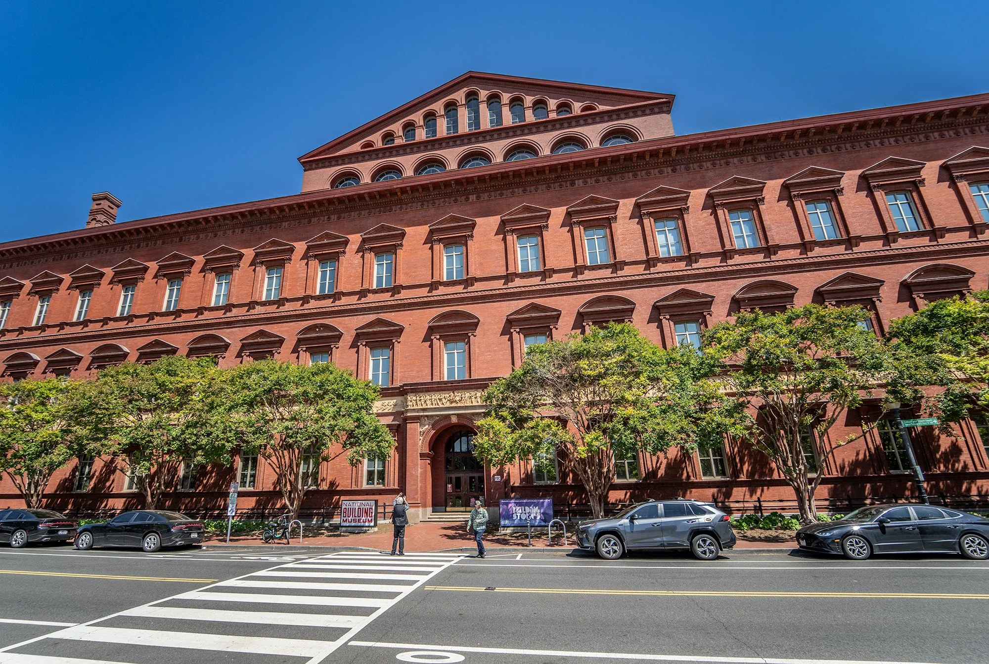 The image shows a historic brick building with multiple windows and decorative architectural details, situated behind a row of green trees. In front of the building, there are parked cars, a crosswalk, and a few people walking or standing on the side