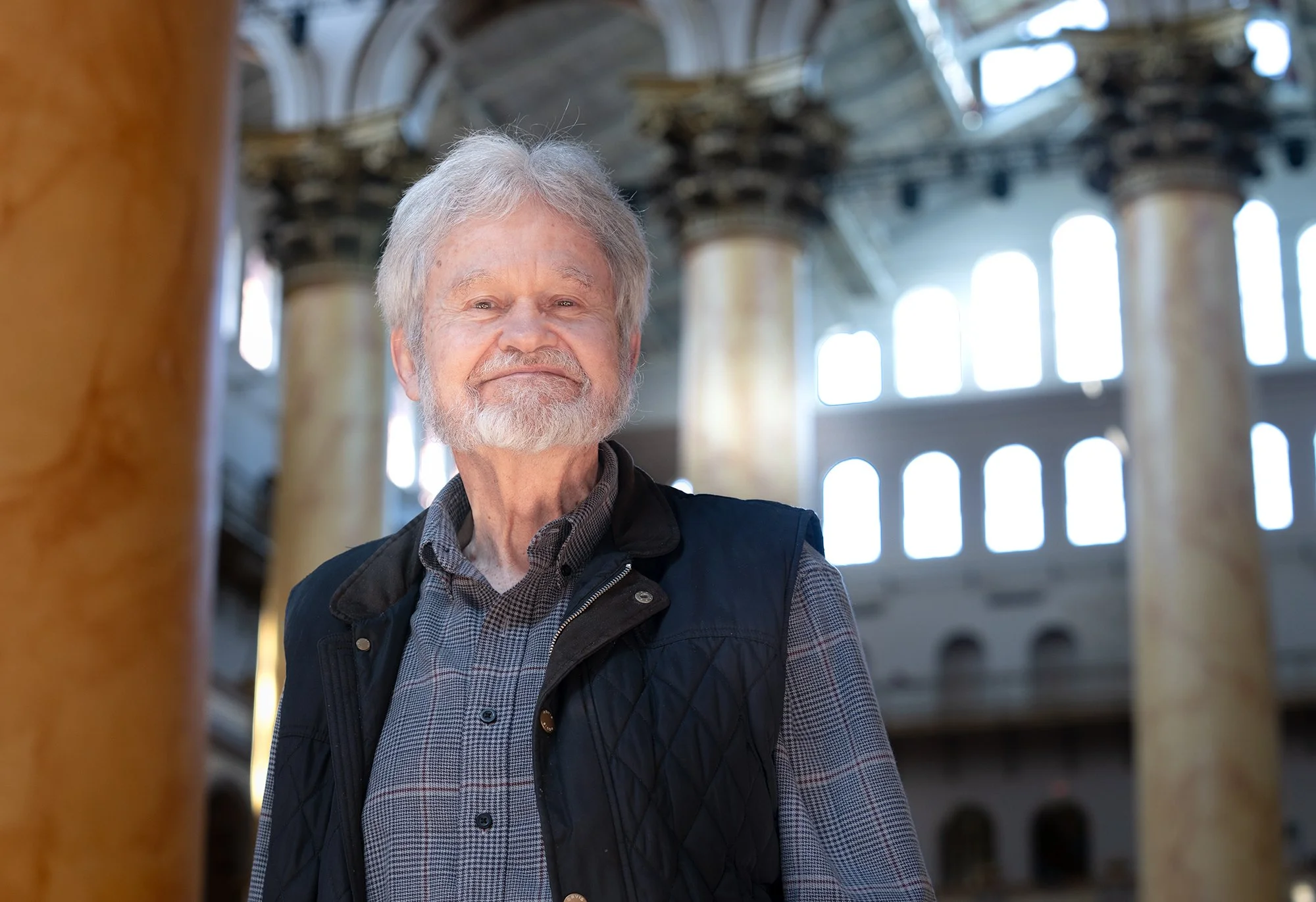 An older man with gray hair and a beard, wearing a checkered shirt and a dark vest, stands inside a building with large windows and grand architectural features.