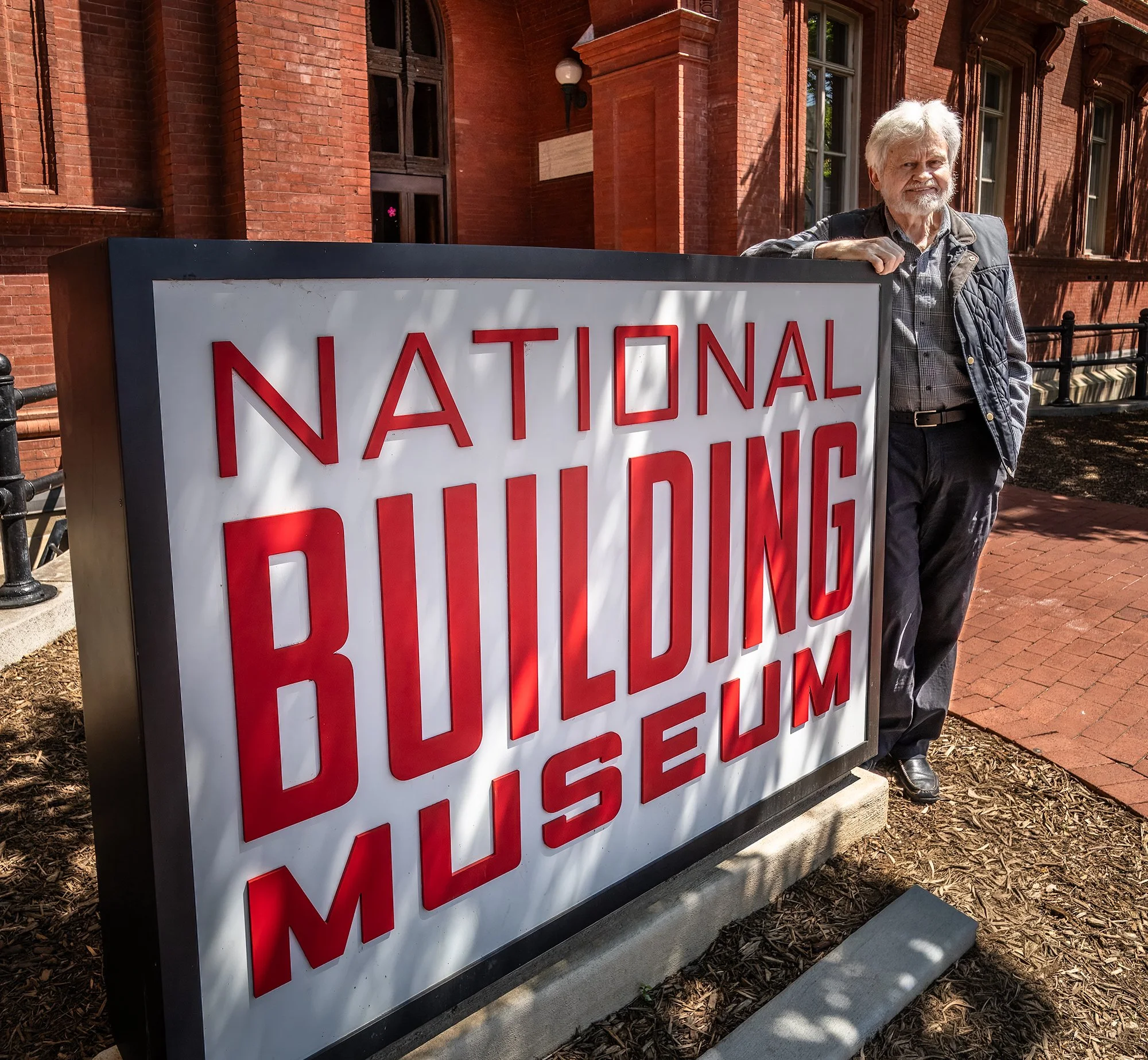 Older man with gray hair and beard standing next to a large white sign with red text outside a red brick building.