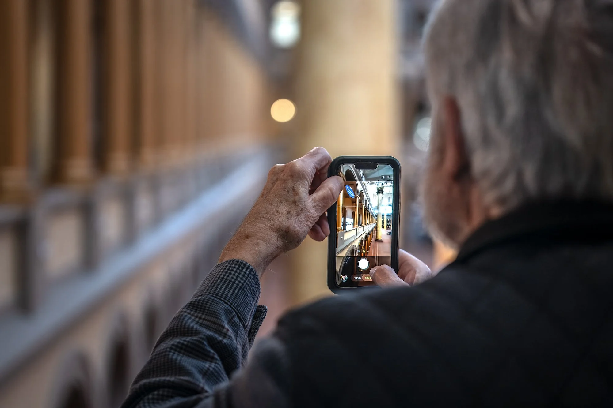 Close-up of an elderly person with gray hair and a checkered blazer taking a photo inside a building with a smartphone.