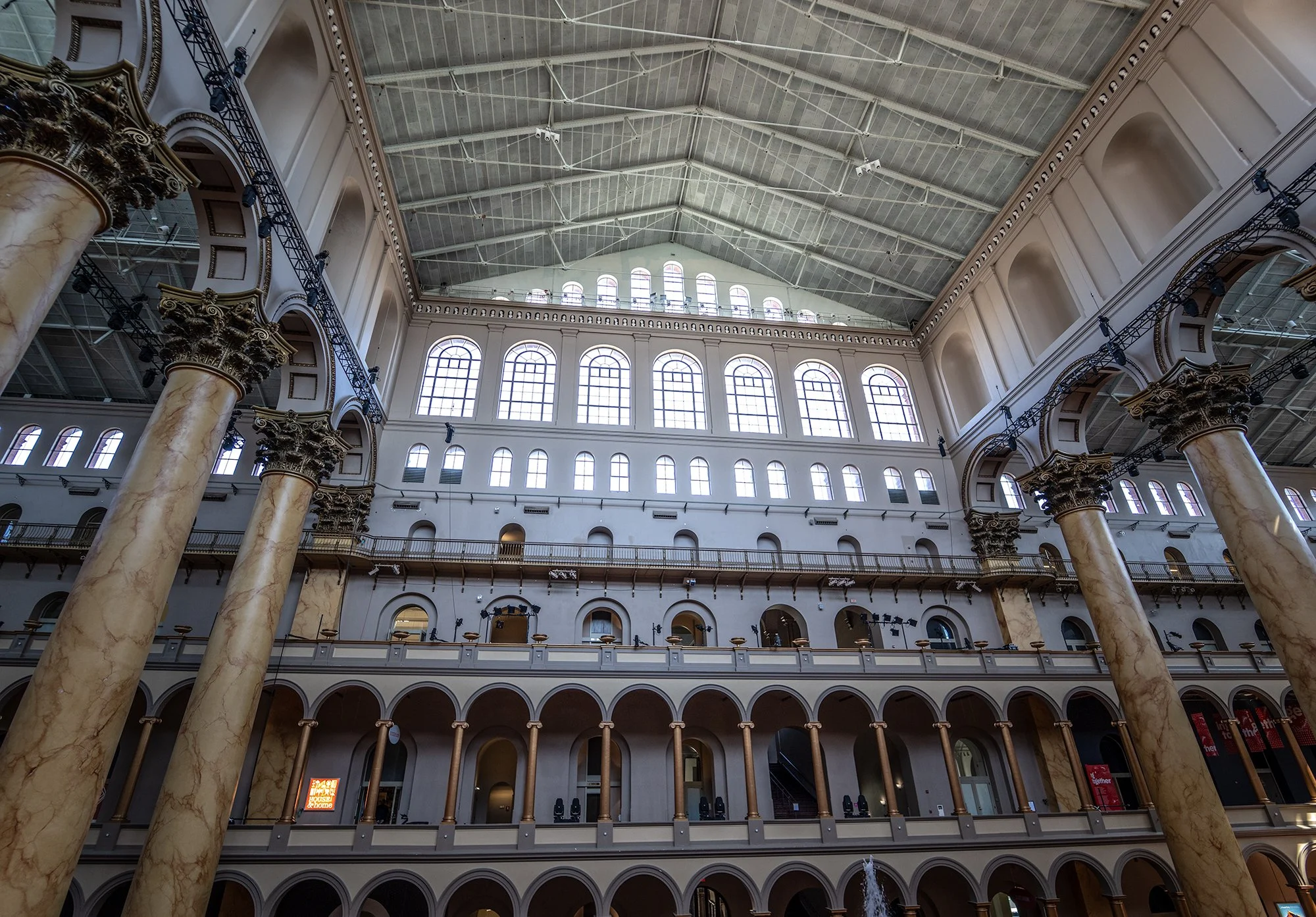 Interior of a large, ornate building with tall marble columns, large arched windows, and a high, vaulted ceiling with visible industrial piping and lighting. There is a balcony or mezzanine level with railings and a large open space beneath.