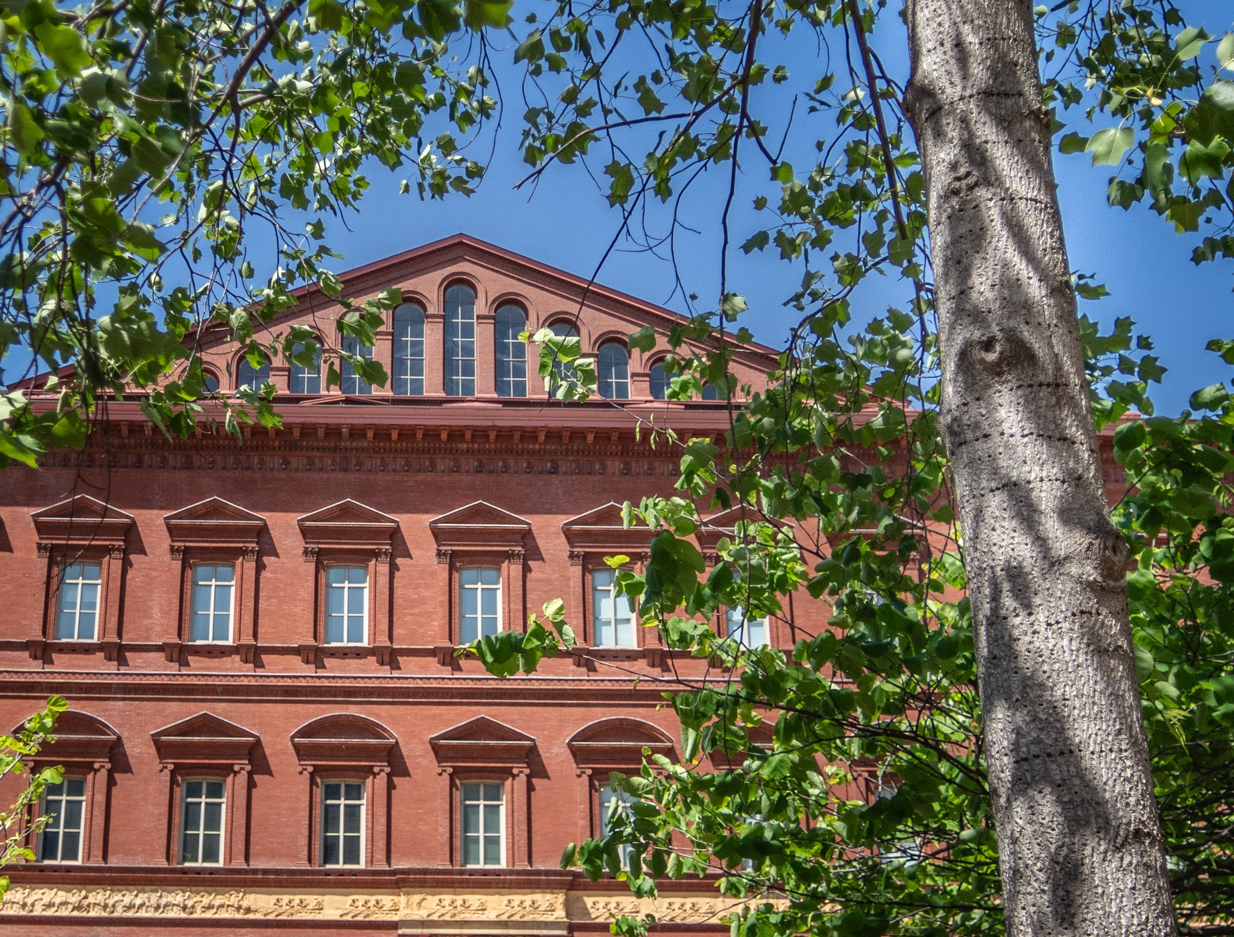 A large brick building with arched windows, seen through green tree leaves and branches, under a clear blue sky.
