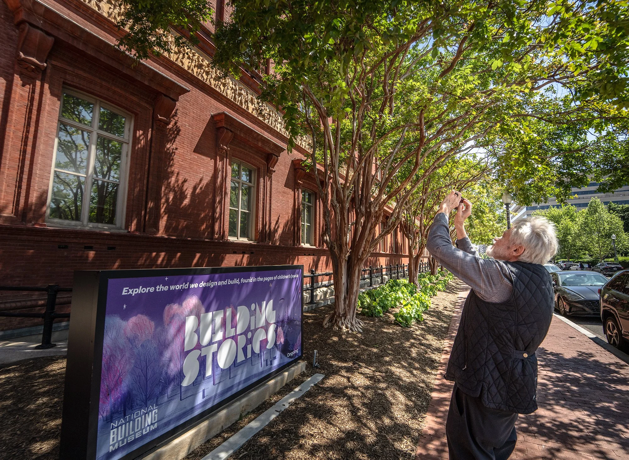 An elderly man with white hair and beard taking a photo of trees outside a brick building with large windows, a decorative frieze, and a painted sign in front of the building.