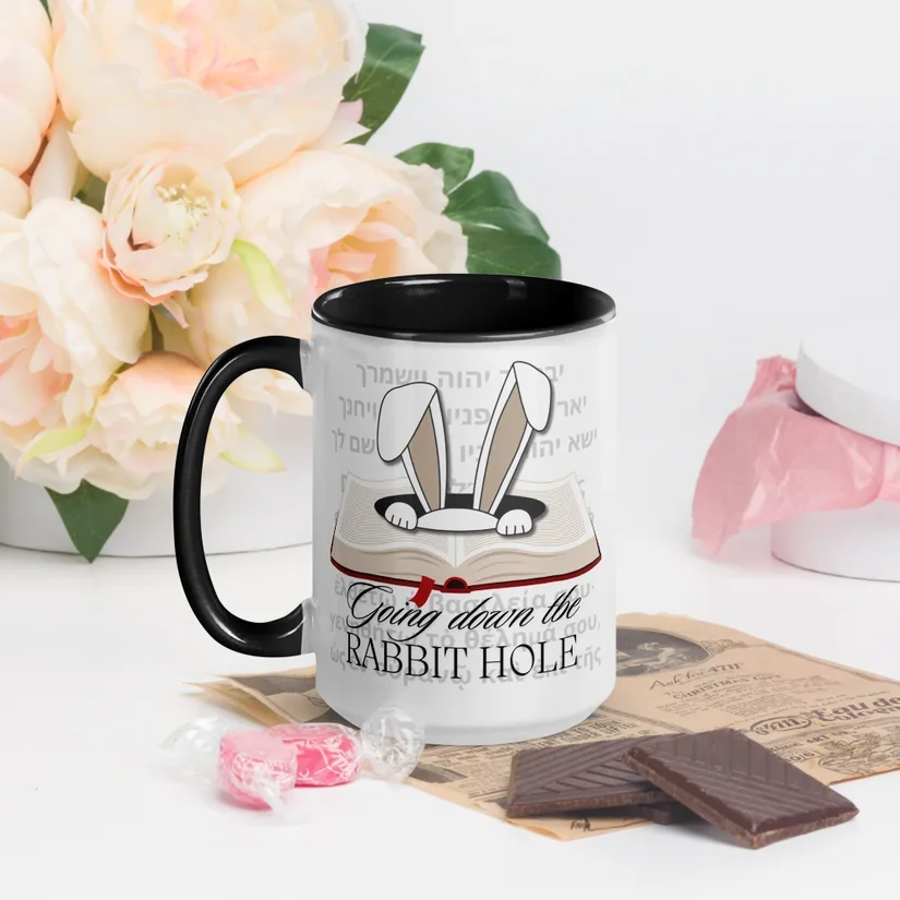 A white mug with a black interior and handle featuring a bunny coming out of a book, with the text "Going down the RABBIT HOLE". The mug is surrounded by pink flowers, wrapped candies, and chocolate on a surface, with pink tissue paper in the background.