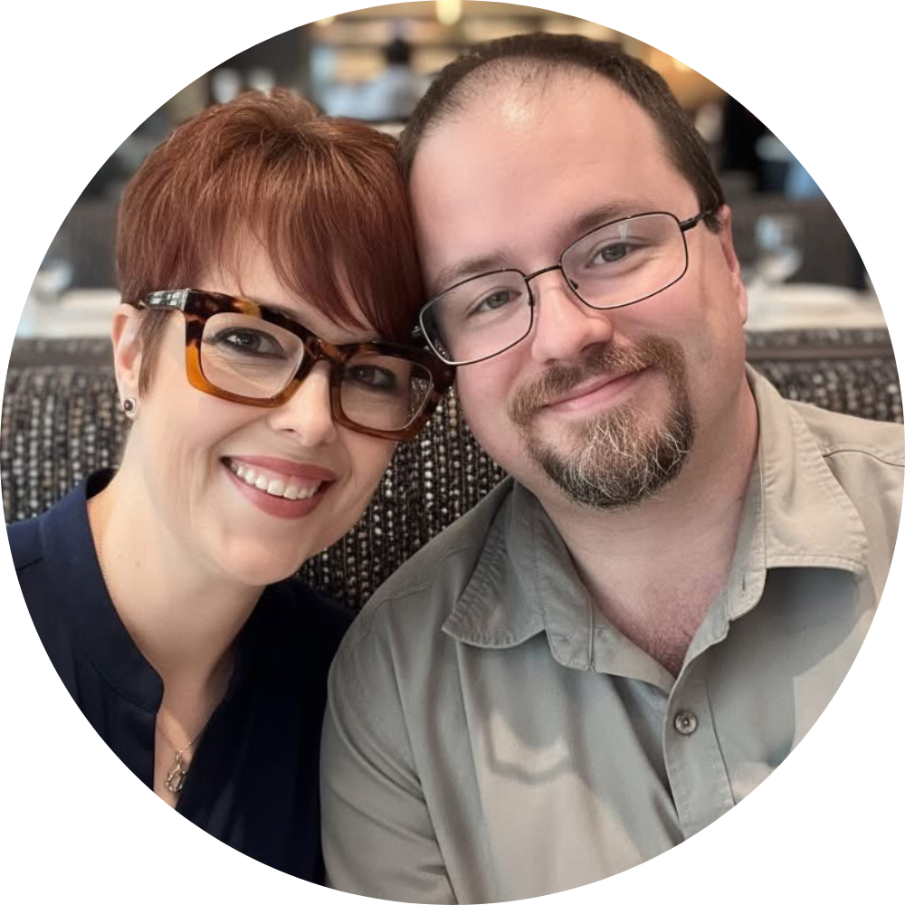 A smiling woman with short red hair, glasses, and earrings, and a smiling man with glasses and facial hair, sitting close together in an indoor setting.