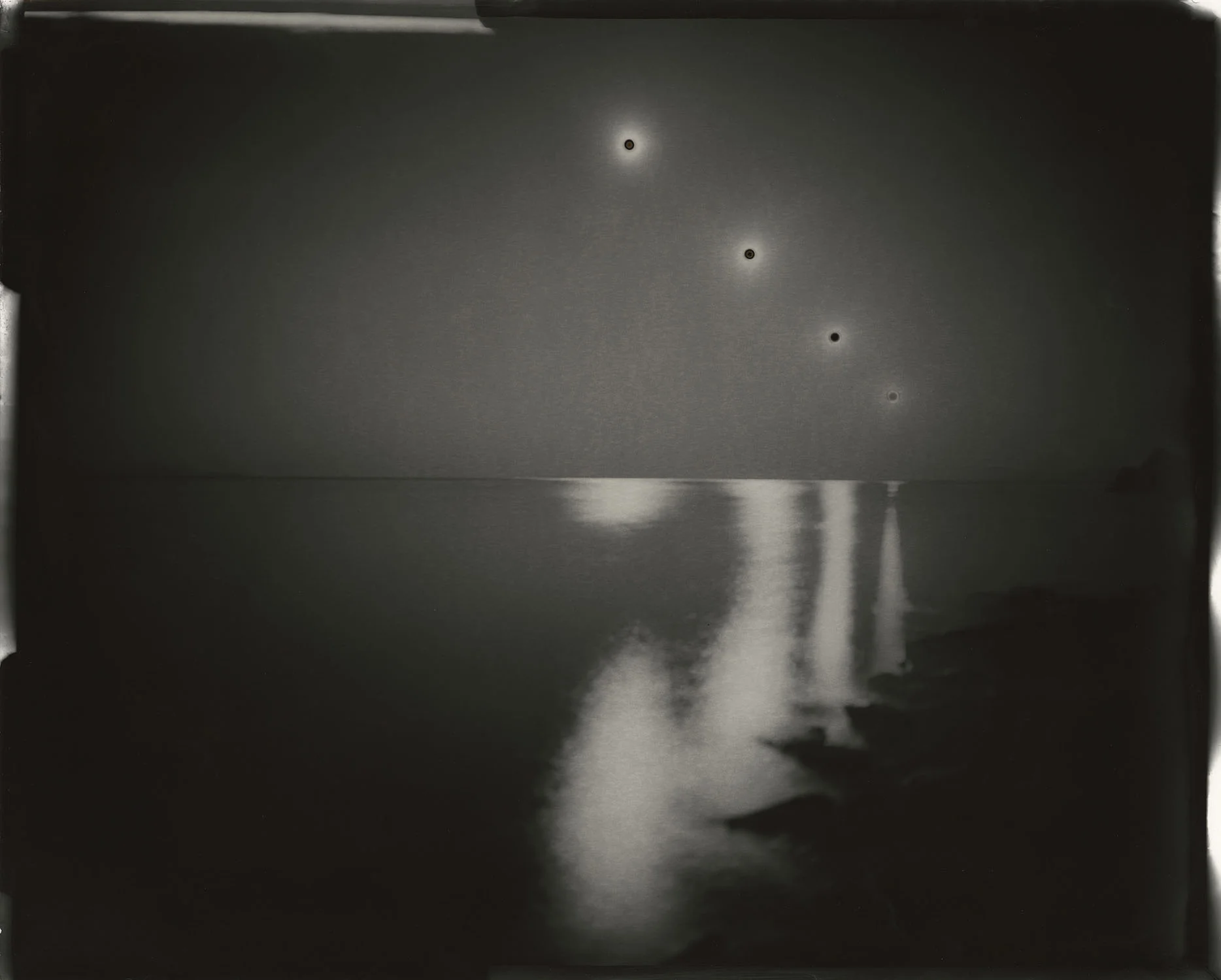Dark monochrome seascape with four bright circular solar marks above the horizon and pale vertical reflections in the water.