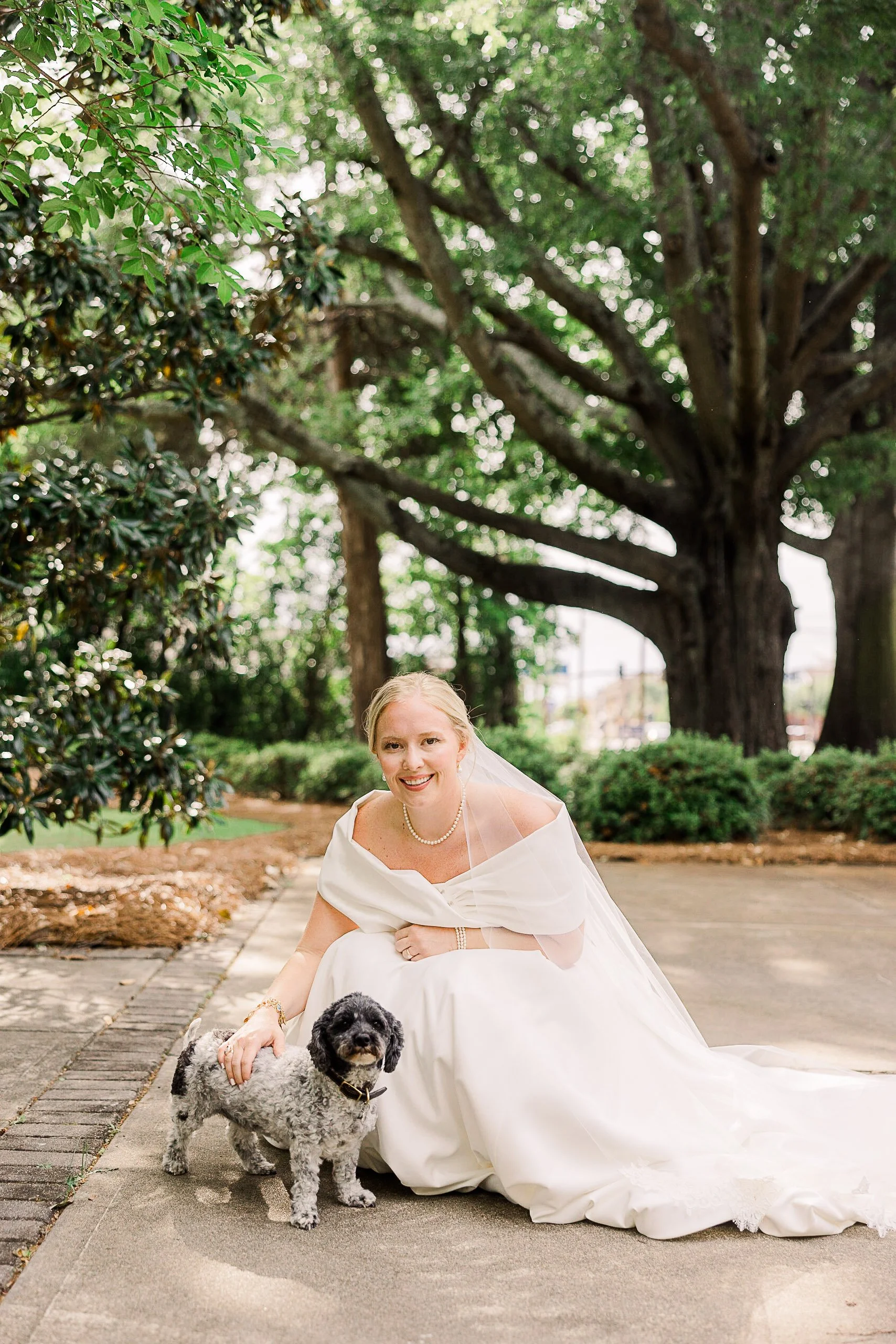 A bride in a white wedding dress kneeling on a sidewalk, smiling at the camera with a black and gray dog, in front of large trees and greenery. Summerville Wedding Photographer