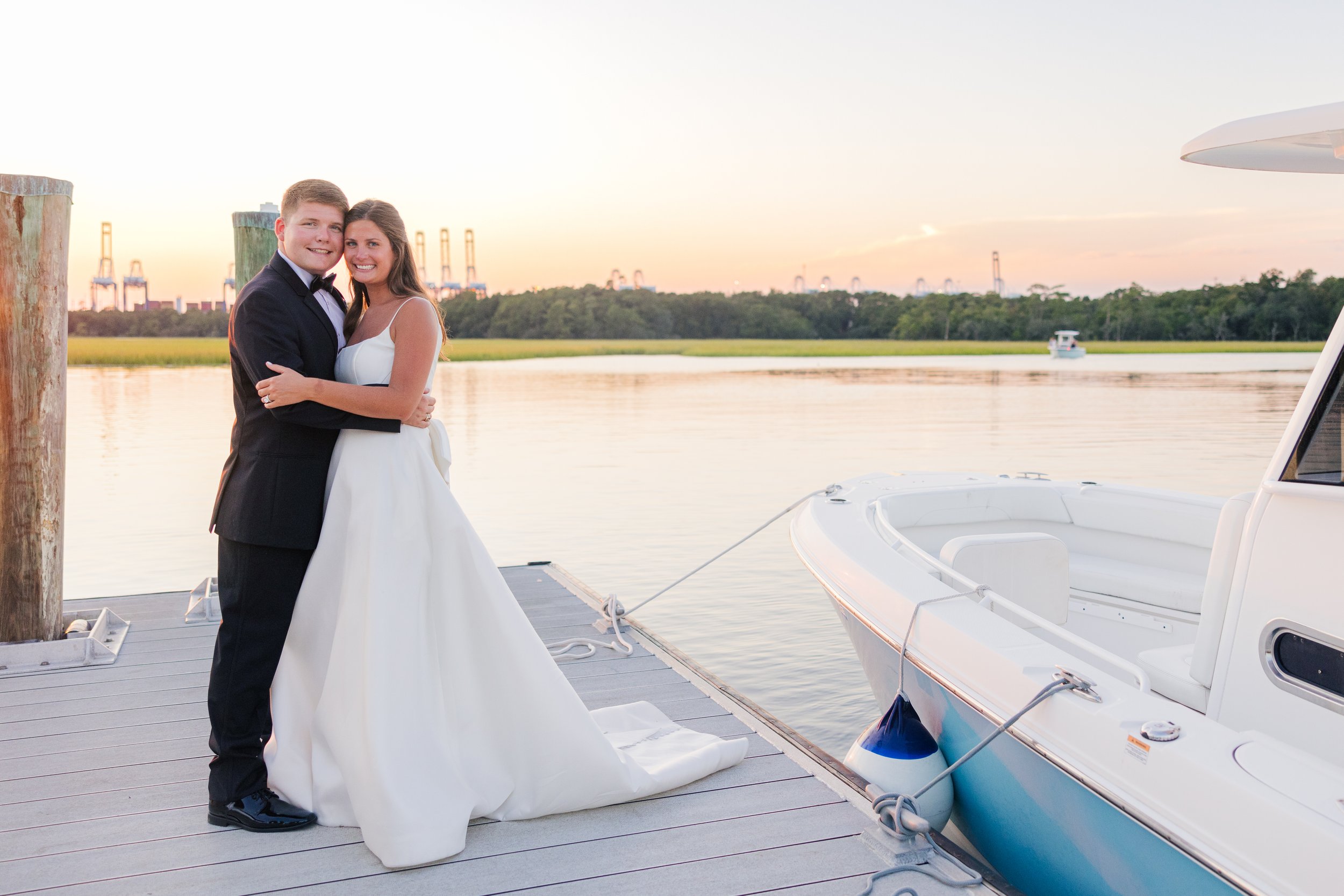 A bride and groom standing close together by a lakeside, holding hands and gazing at each other, under large trees with overhanging branches, during sunset. Natural Wedding Photographer