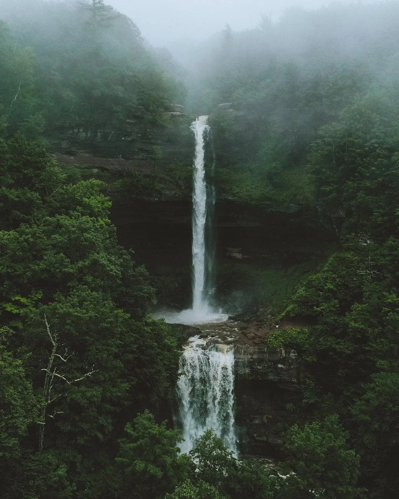 Tall waterfall cascading down a rocky cliff surrounded by lush green forest and mist.