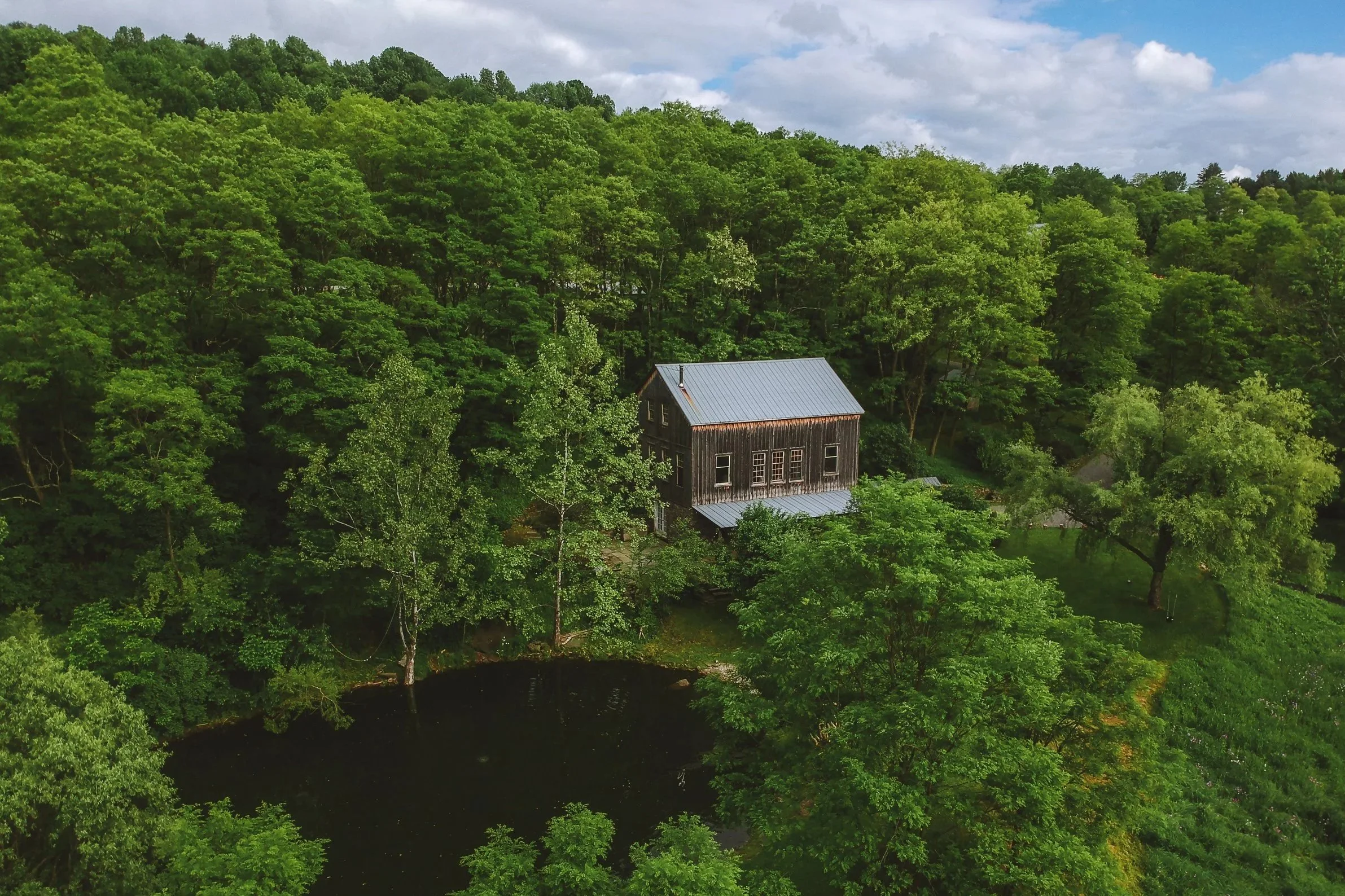 A wooden house with a metal roof is surrounded by lush green trees and hills, with a small pond nearby under a partly cloudy sky.