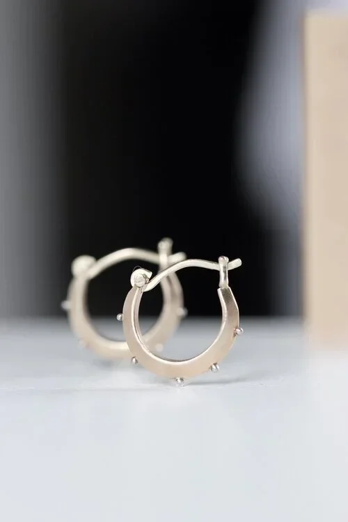 Silver hoop earrings with small decorative beads placed on a flat surface with a blurred dark background.