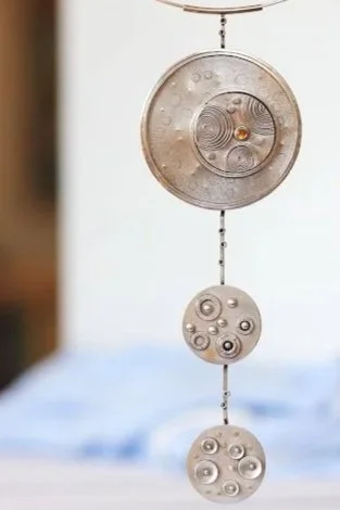 A kinetic metal sculpture with three circular discs connected by a chain, hanging from a metal hook, with a blurred background.