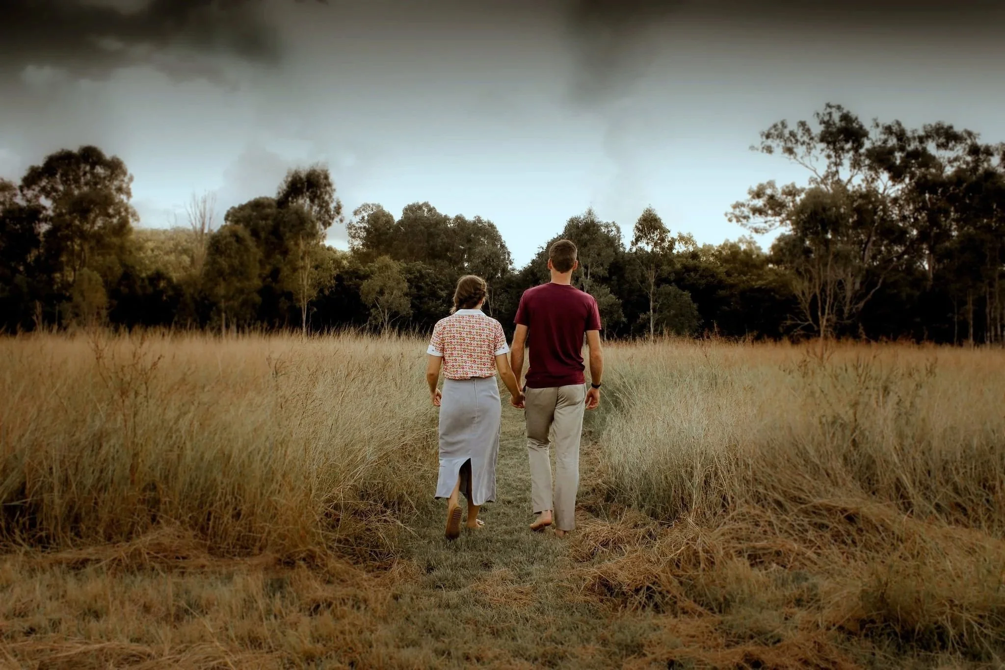 A couple walking hand in hand along a grassy path in a field with tall golden grass, with a backdrop of trees and a cloudy sky.