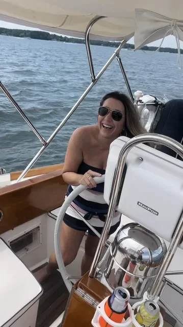 A woman on a boat smiling and holding the steering wheel, wearing sunglasses with water and shoreline in the background.