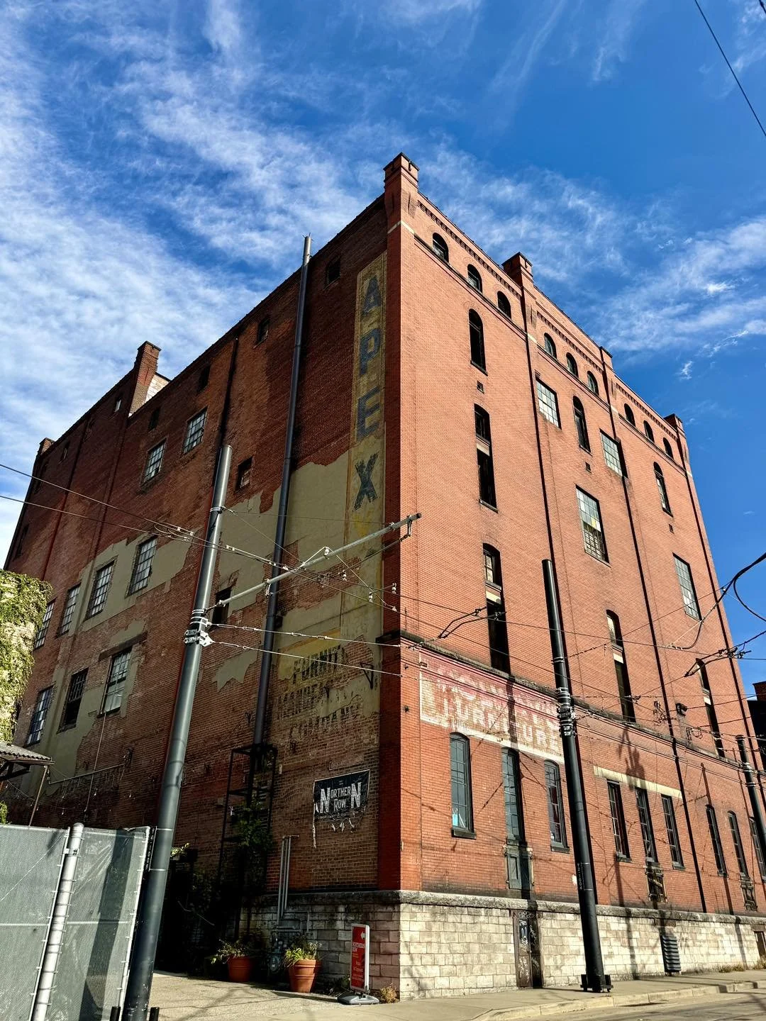 Red brick multi-story building with vintage painted advertising signs on its side, set against a blue sky with wispy clouds. Urban street scene with power lines and sidewalk.