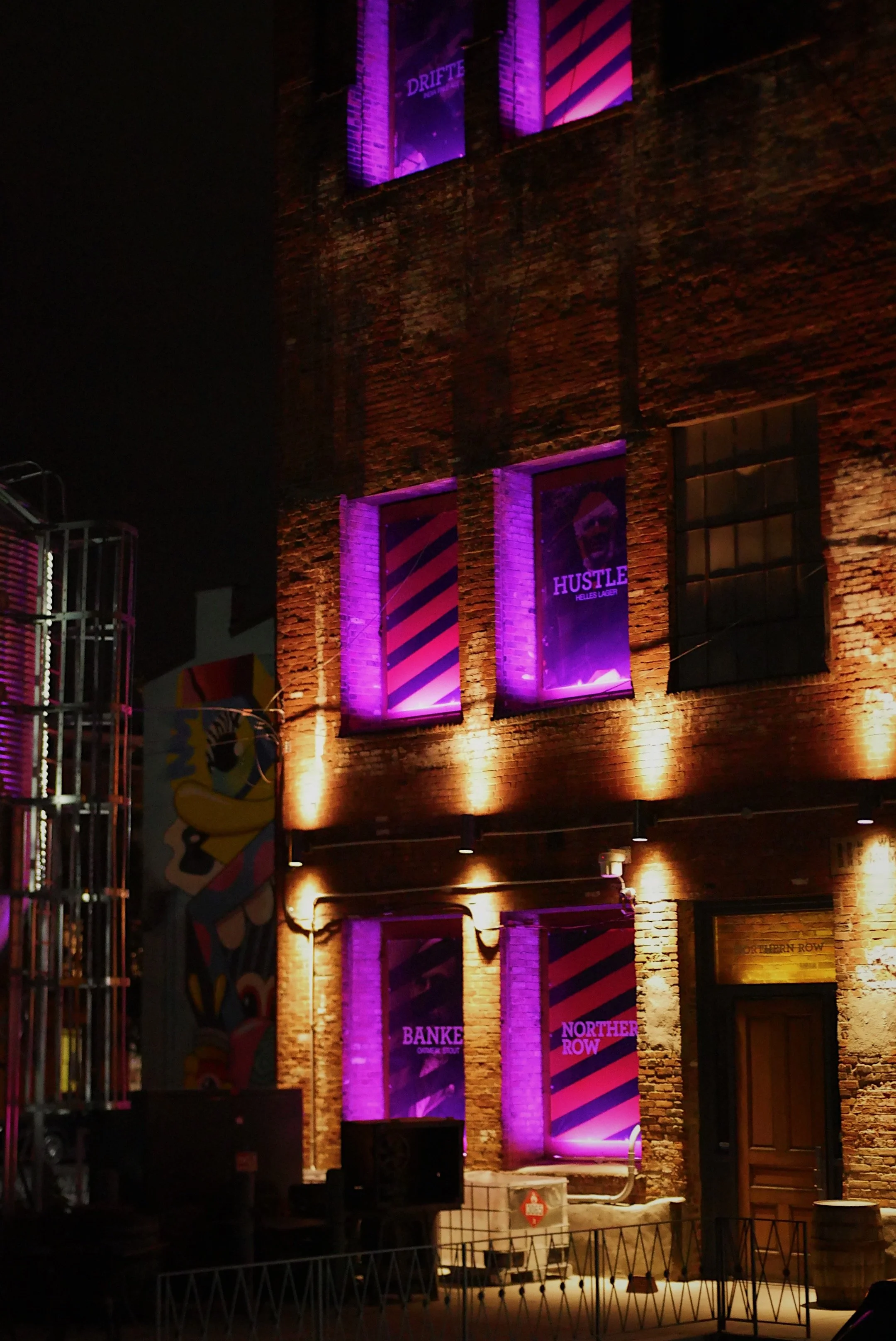 Night view of a brick building with illuminated purple banners on its windows. The banners display words like "Hustle", "Bank", and "Northern Row". The building is lit with warm lights at the ground level.