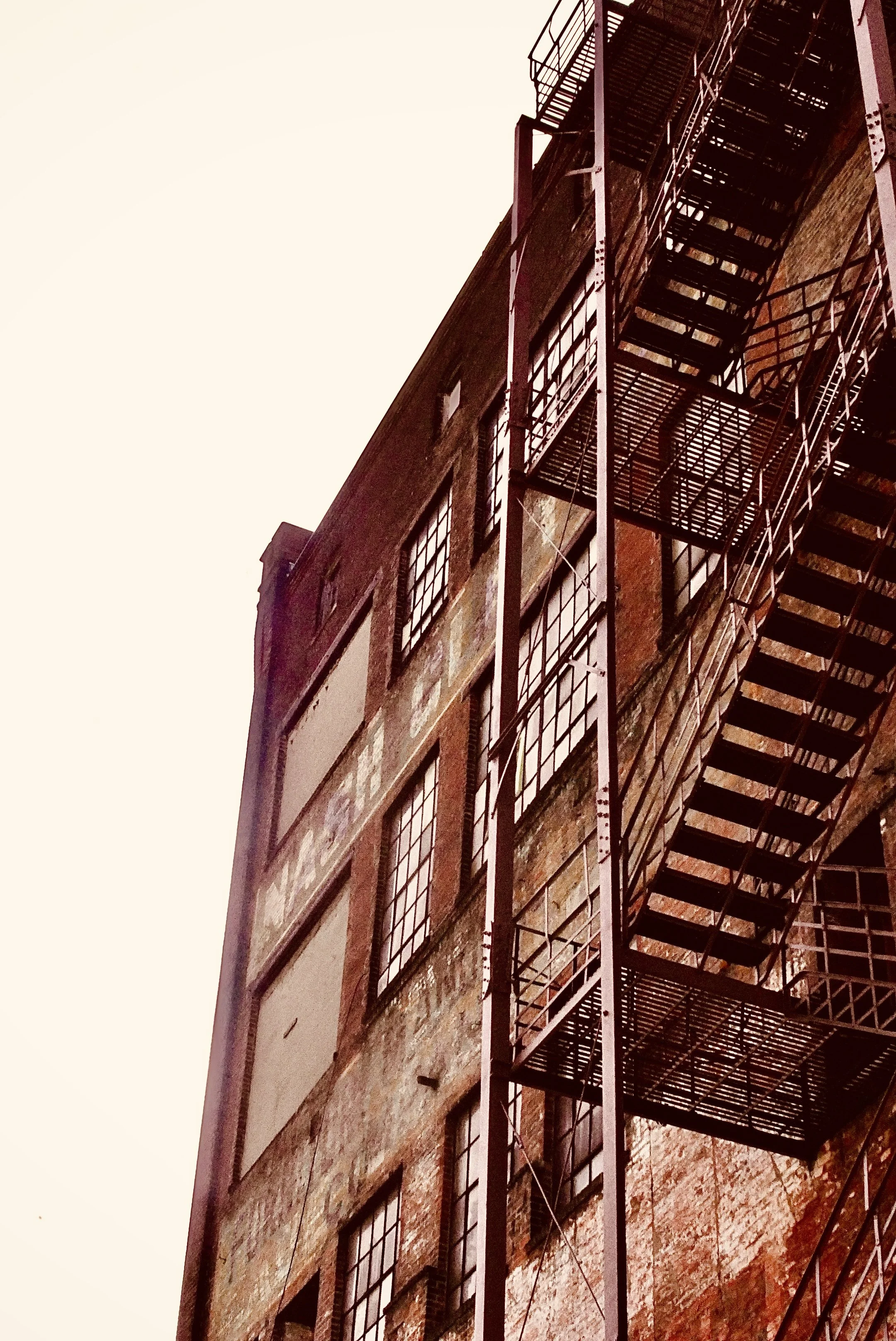 A tall brick building with fire escape stairs and multiple windows, viewed from below against a pale sky.