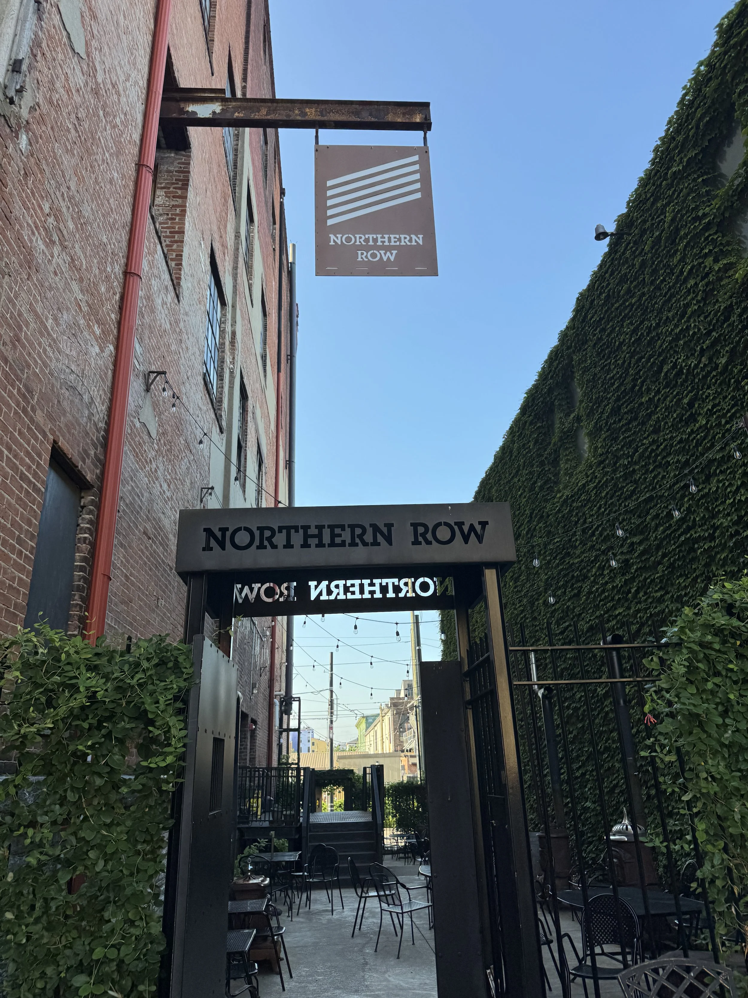 Entrance to Northern Row with outdoor seating and string lights, brick building on the left and greenery on the right, clear blue sky.