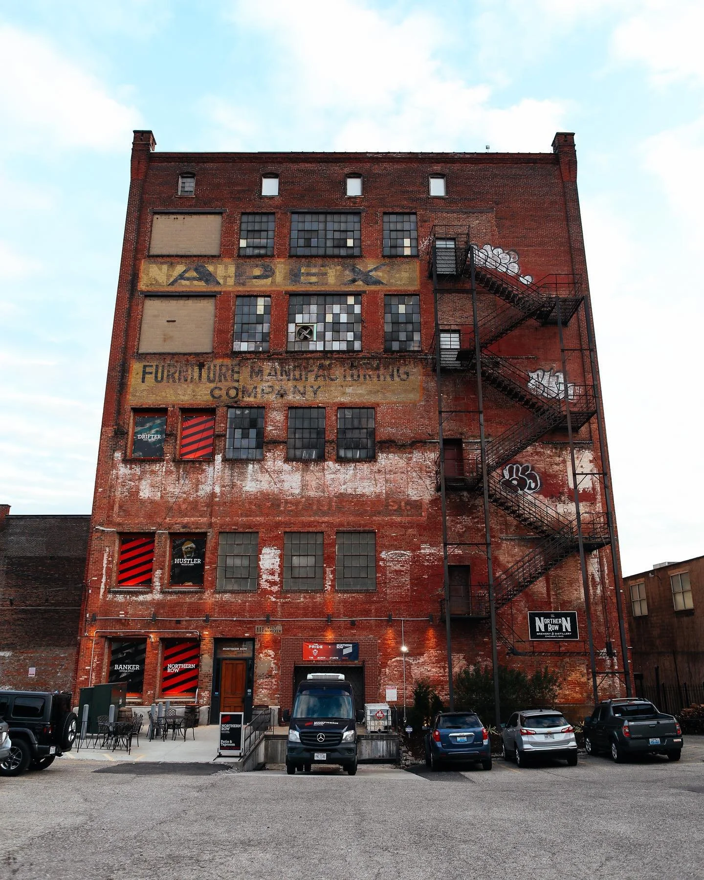 A tall, red brick building with multiple windows and some broken or boarded-up sections. There is an old faded sign on the building that reads 'NORDX FURNITURE MANUFACTURING COMPANY'. A black fire escape runs down the right side of the building. Seve