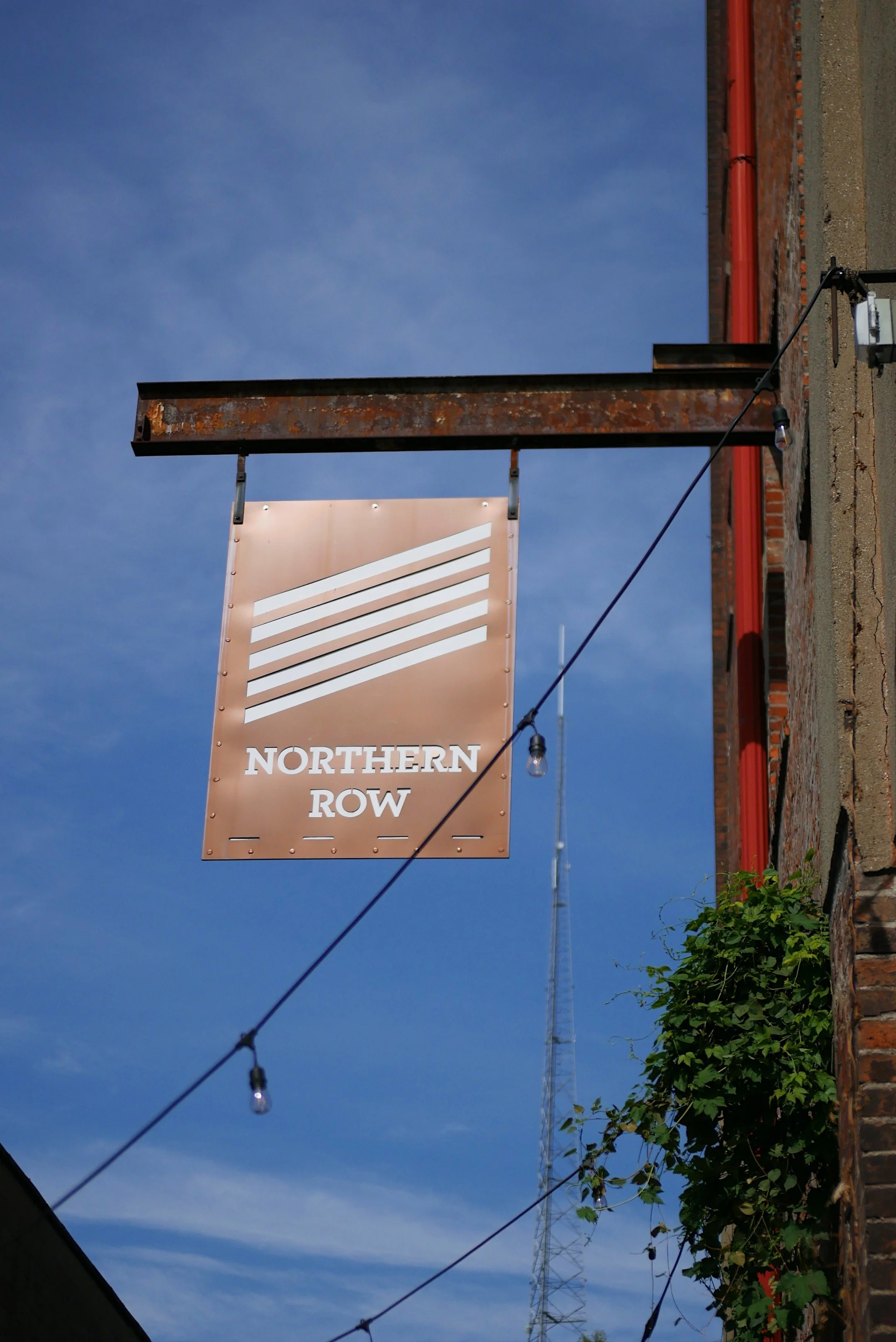 A hanging sign for Northern Row with a logo of three diagonal stripes, located on the side of a brick building under strings of small hanging lights, against a blue sky background.