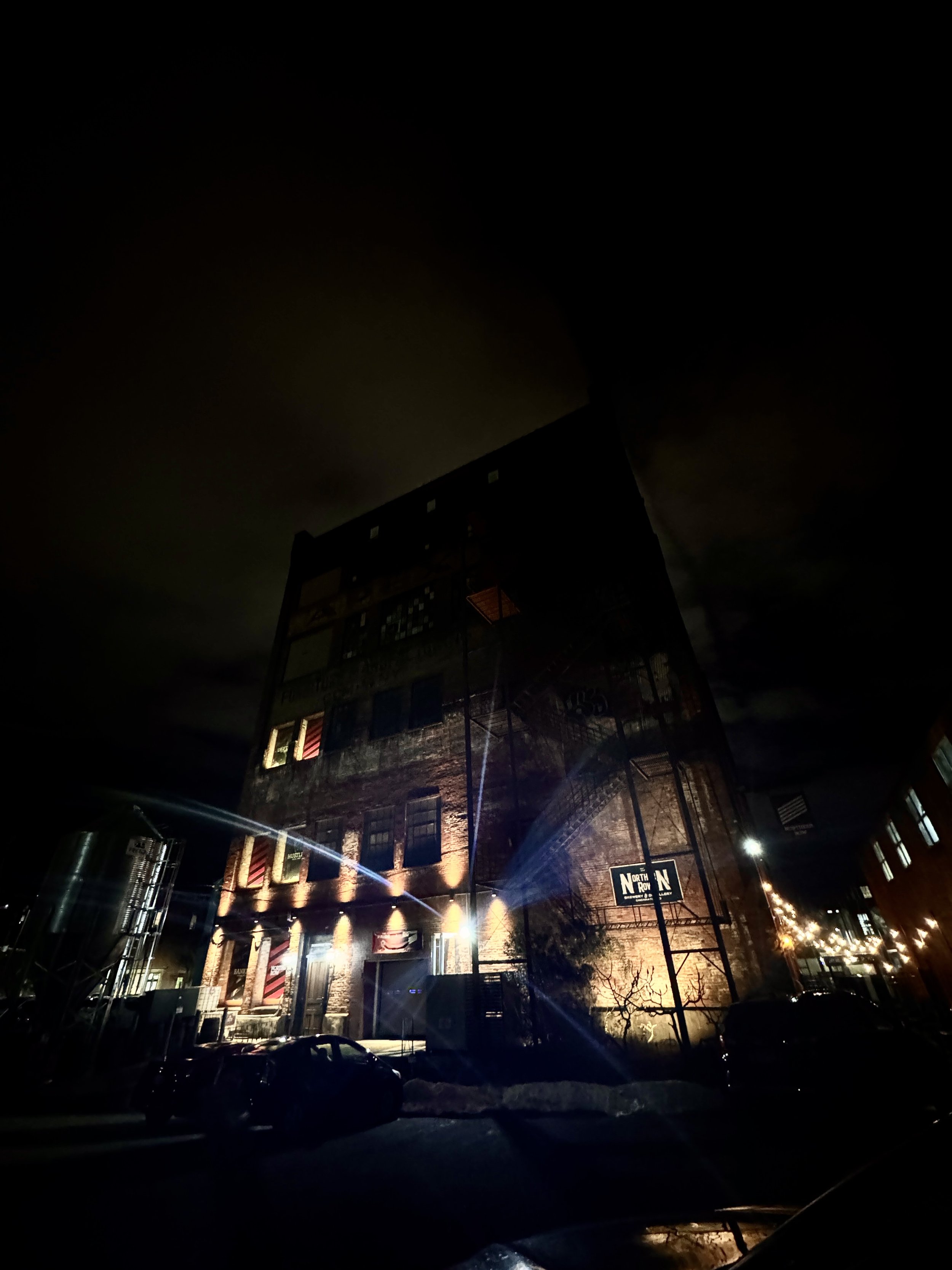 Nighttime view of a tall brick building illuminated by exterior lights, with a few visible windows and parked cars in the foreground, and cloudy sky above.