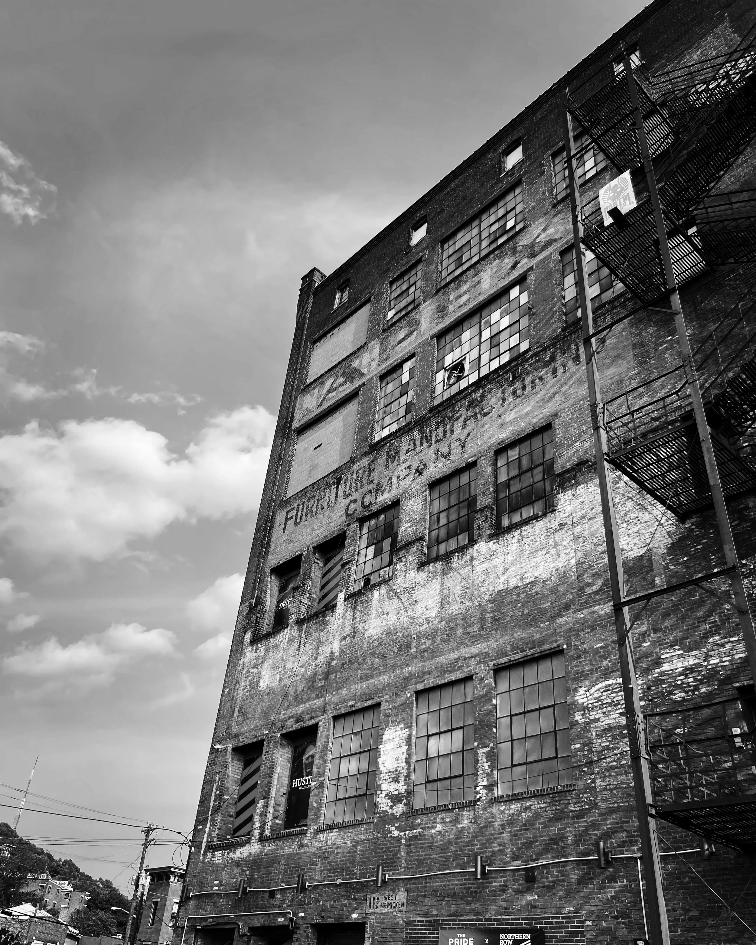 A tall, old brick building with multiple large windows and an exterior fire escape. The building has faded painted signage on its side, and the sky above is partly cloudy.