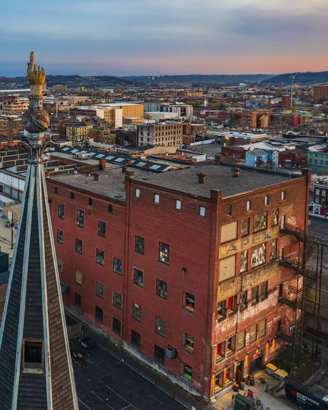 Aerial view of a city skyline at sunset, featuring an old brick building with fire escape stairs, a spire with a hand sculpture, and numerous other buildings in the background.
