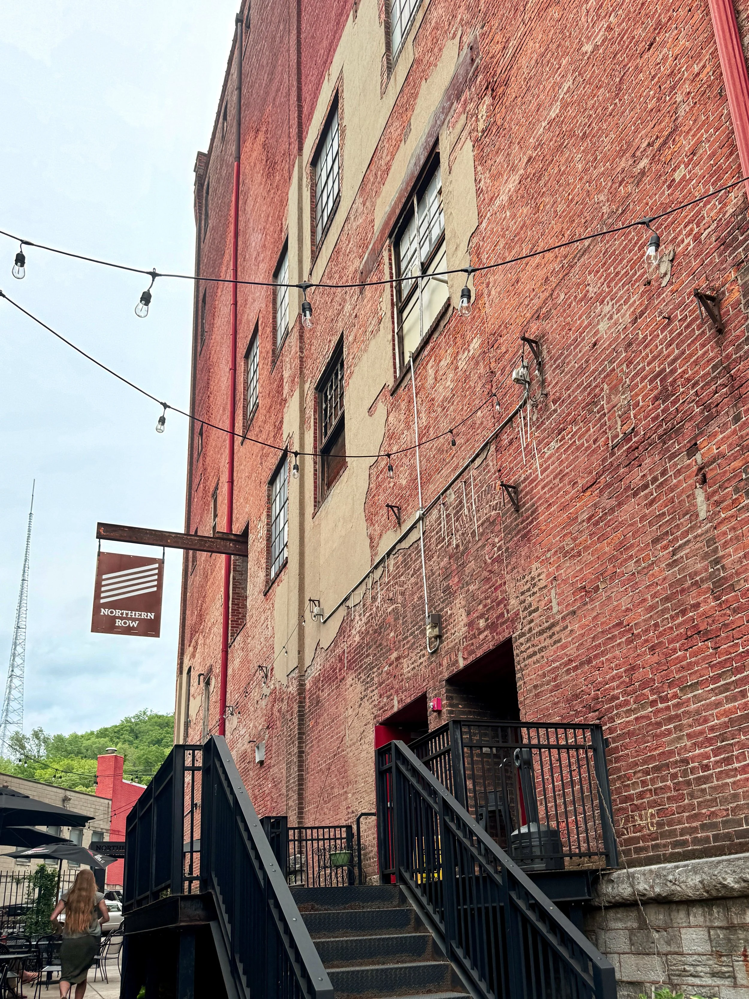Outdoor scene of a red brick building with a staircase leading up to an entrance. String lights are hanging across the view. There is a sign that says 'Northern Row'. A woman with long blonde hair and a green dress is walking away near outdoor seatin