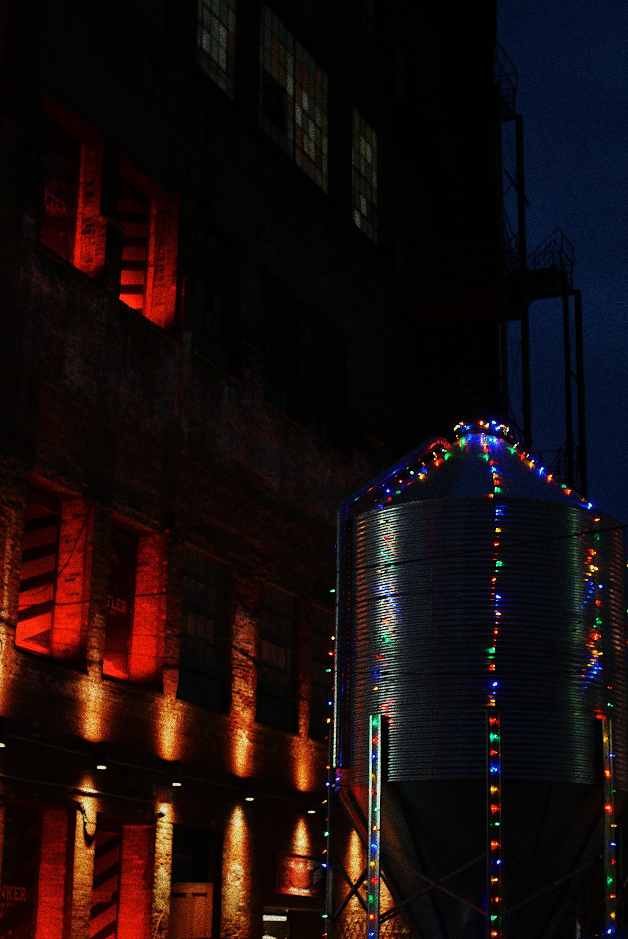 Nighttime scene with a large metal silo decorated with colorful Christmas lights, nearby a brick building with illuminated red windows.