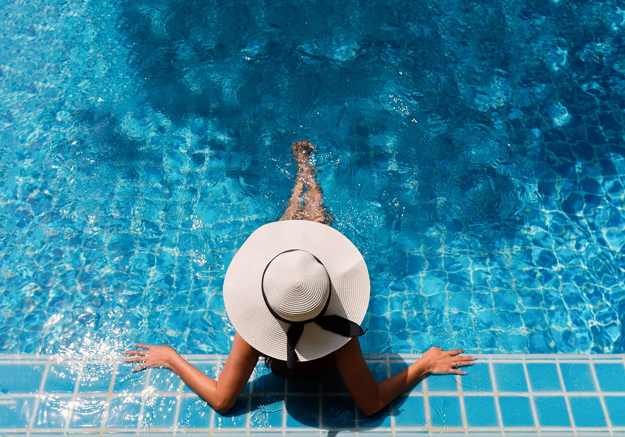 A woman in a black swimsuit and a wide-brimmed straw hat with a black ribbon is sitting on the edge of a swimming pool, with her feet in the water and her arms resting on the pool edge. The pool has blue tiles and the water is clear and blue.