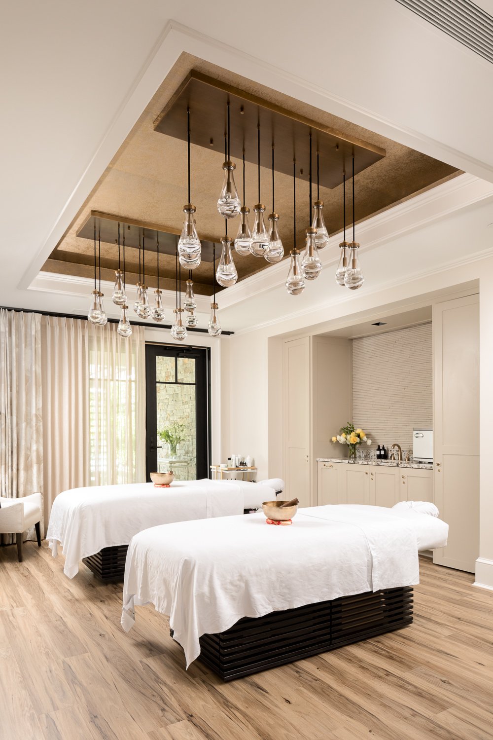 A luxurious spa or massage room with two massage tables draped in white sheets, wooden bowls on each table, elegant glass pendant lights hanging from a recessed ceiling, large windows with sheer curtains, and a small kitchenette with flowers and skin