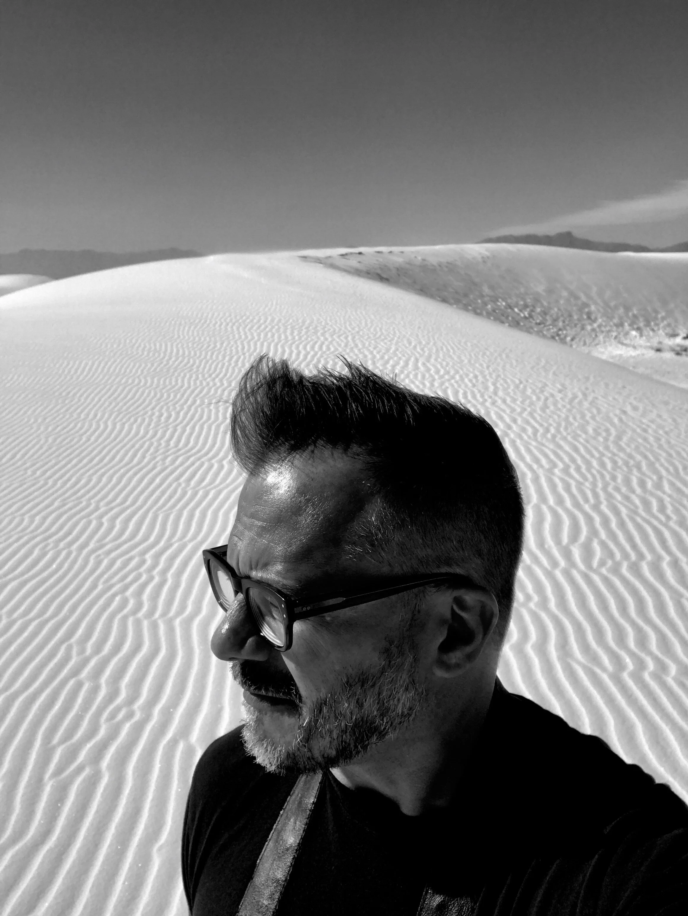 A man with glasses and a beard, wearing a cap, with a backpack strap, in a snowy desert landscape with sand dunes and mountains in the background.