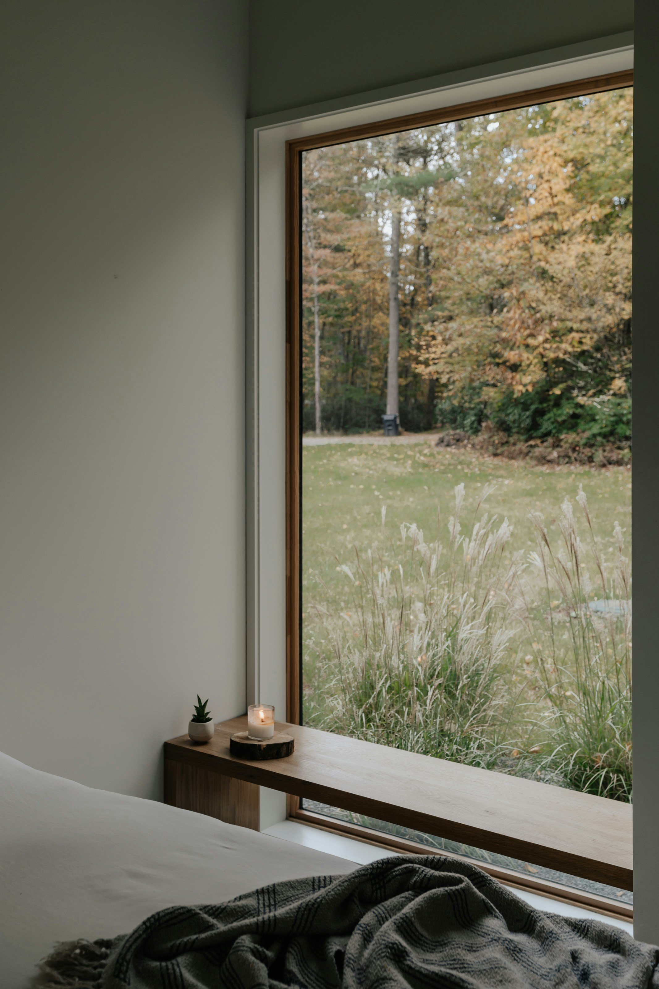 A bedroom with a large window showing an outdoor scene of trees with autumn-colored leaves. Inside, a wooden surface with a small potted plant and a lit candle is visible near the window, with a bed in the foreground covered with a gray wool blanket.