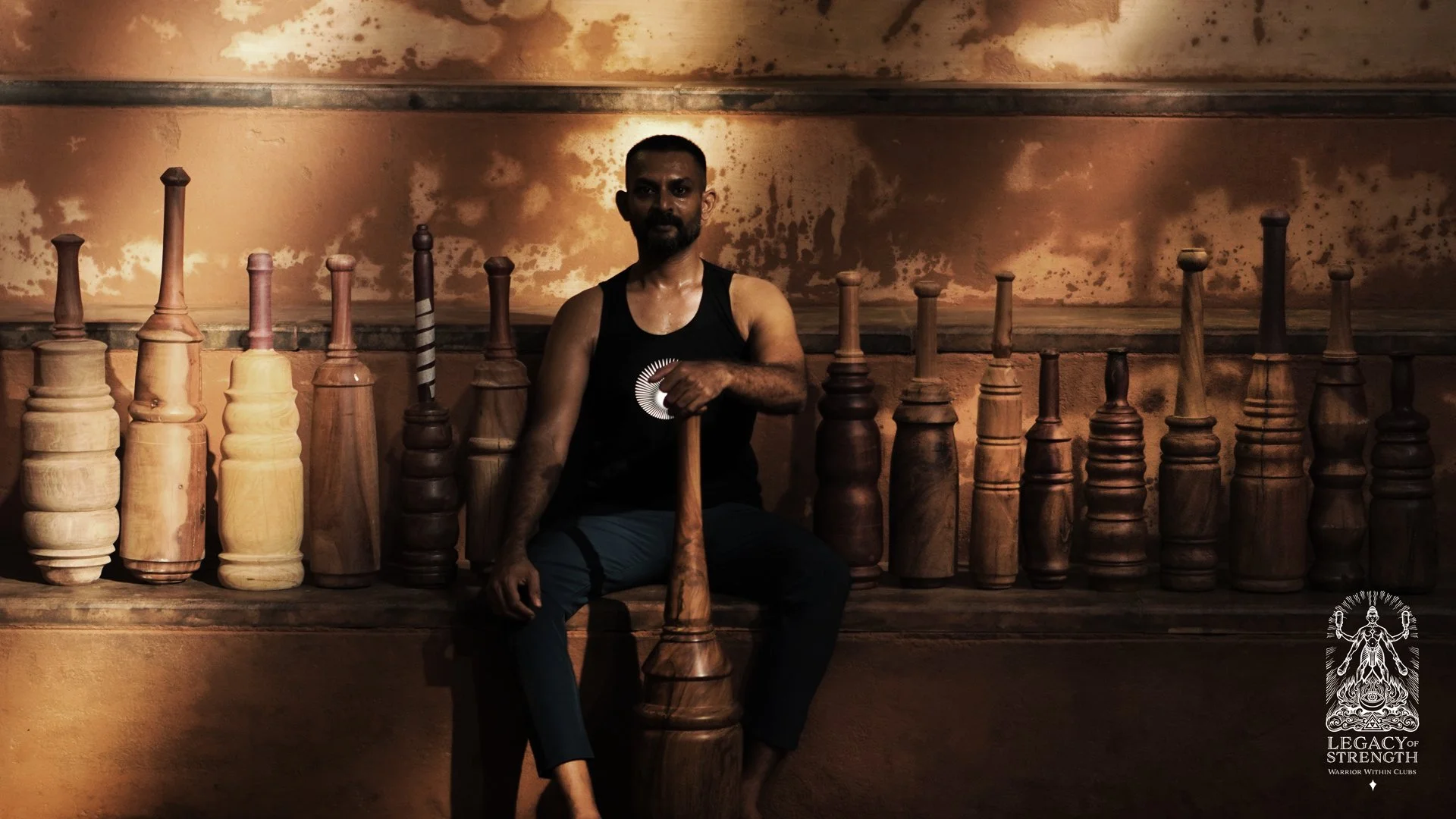 A man sits among wooden chess pieces in a dimly lit room with a warm, textured background, wearing a black tank top and blue pants, holding a large wooden club vertically.