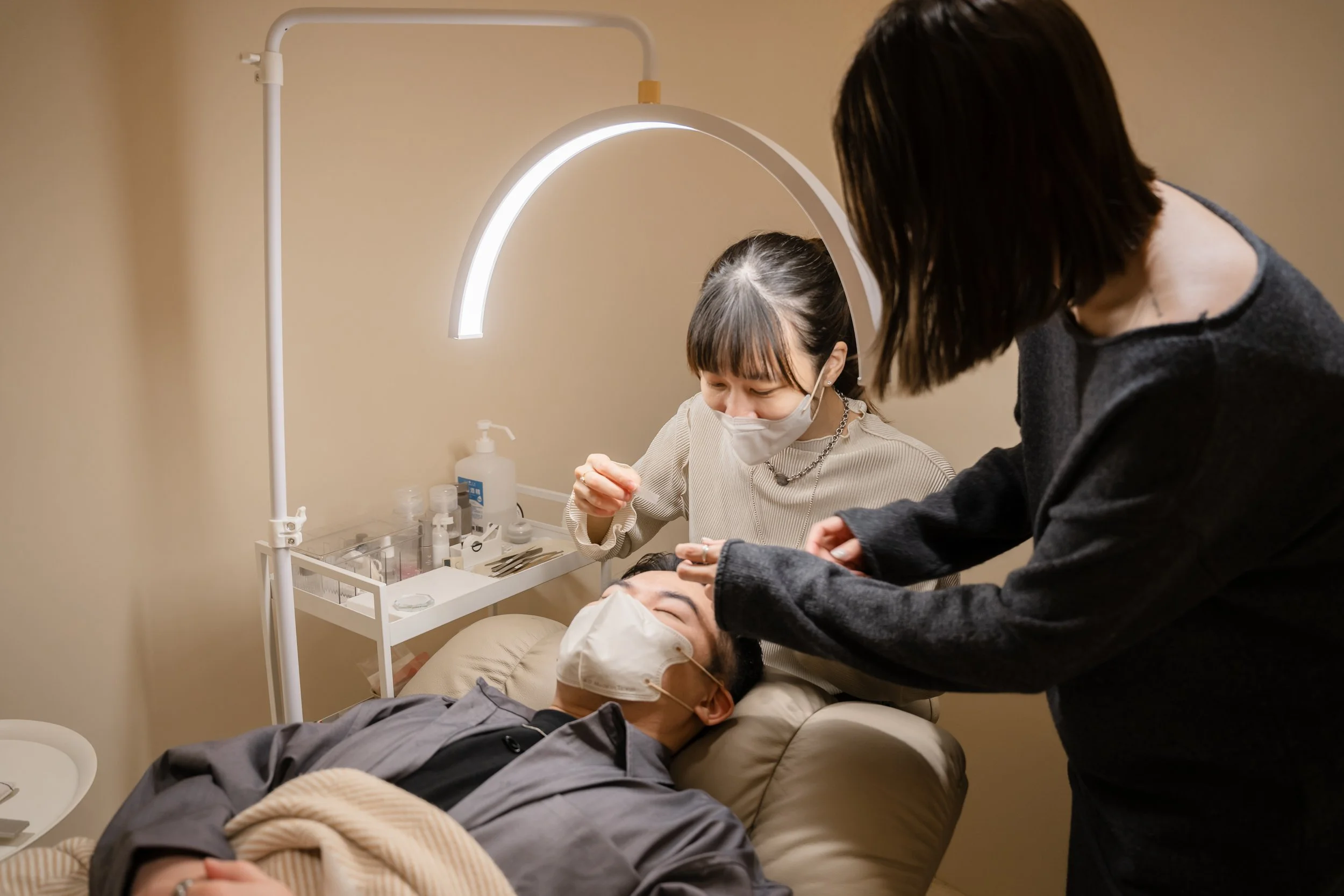 A person lying on a reclined chair receives a cosmetic or medical treatment from two women wearing masks indoors, with a small table of supplies nearby.