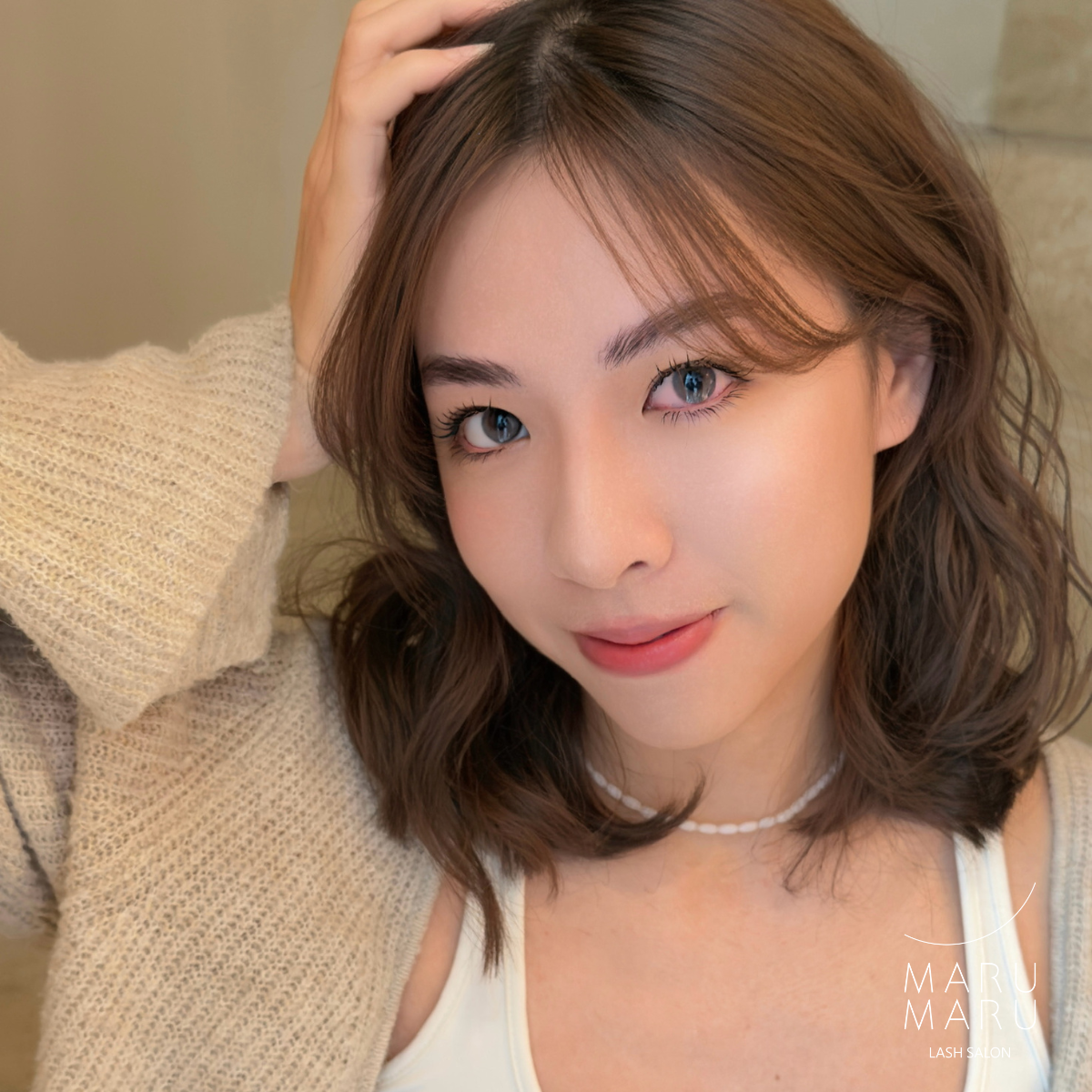 A woman with wavy brown hair, blue eyes, and light skin tone, wearing a beige knitted sweater and a white tank top, smiling and touching her hair, with a blurred background.