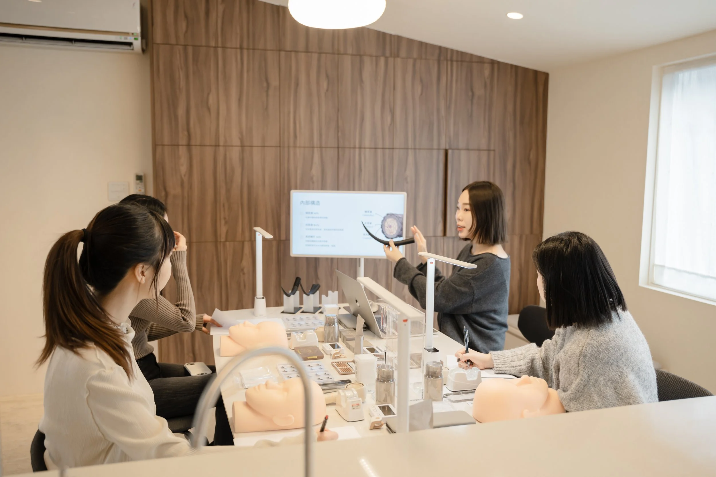 A woman instructor is giving a skincare or cosmetic training class to four students in a room with wooden paneling. The instructor points at a presentation slide on a screen, showing skin or facial anatomy. The students are seated at a table with skincare training models, notebooks, and pens.