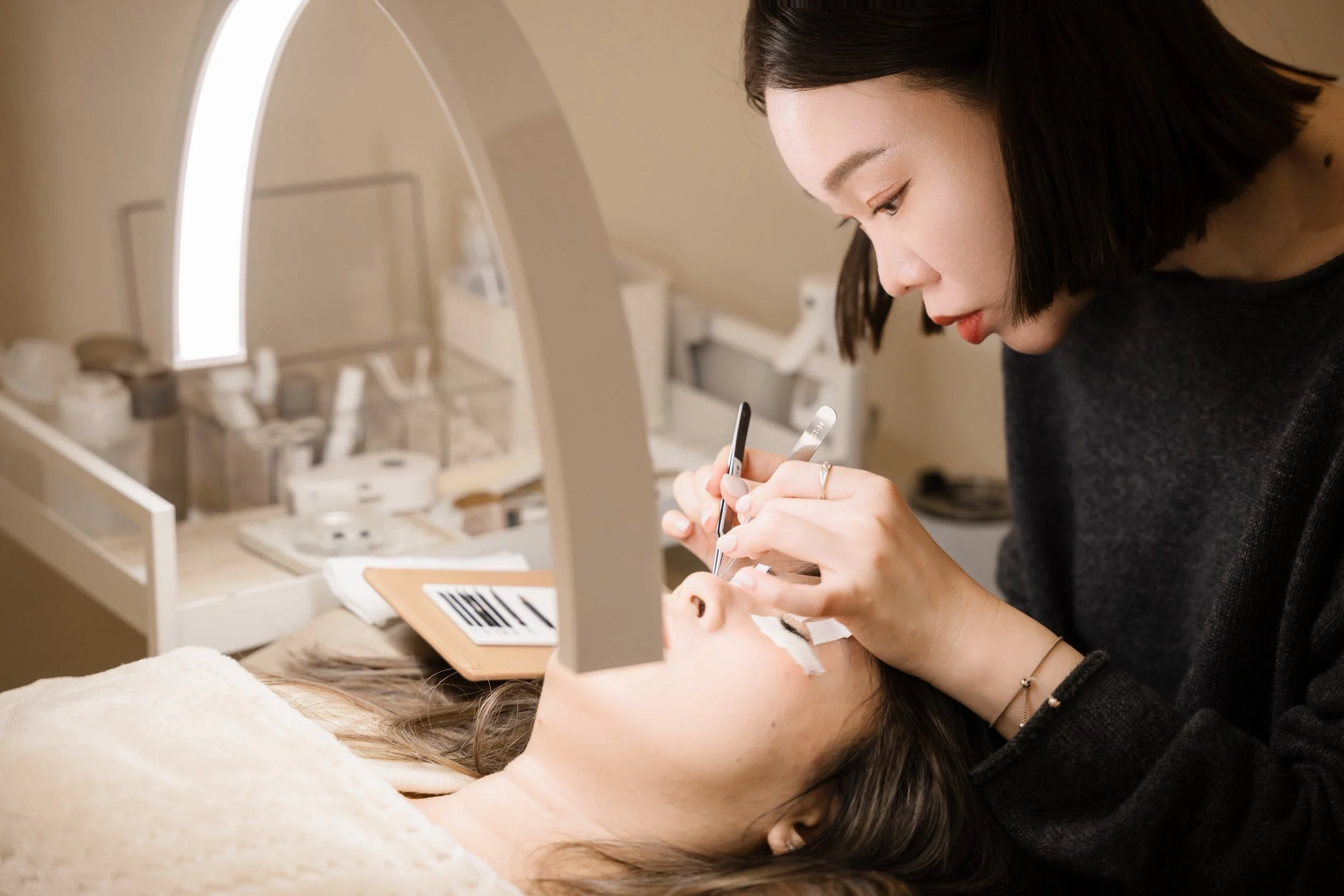 A woman having a makeup procedure done by a makeup artist, who is holding makeup brushes and tools near her face in a clinical setting.