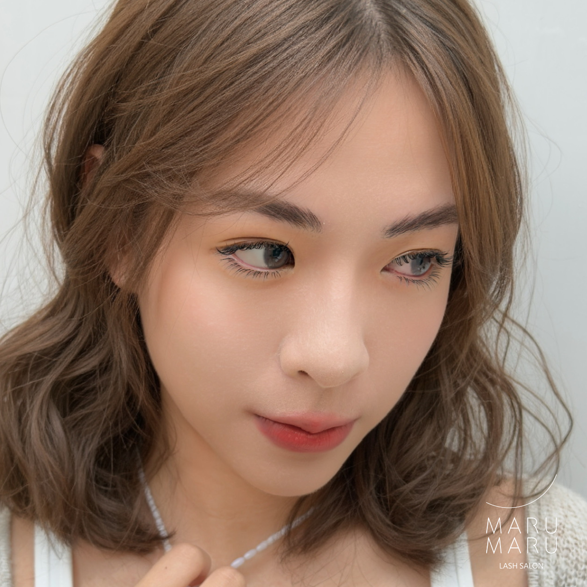 Close-up of a young woman with wavy brown hair, light makeup, and a nude lipstick, looking slightly downward with a neutral expression. She is wearing a white top and a thin necklace, with a blurred white background.