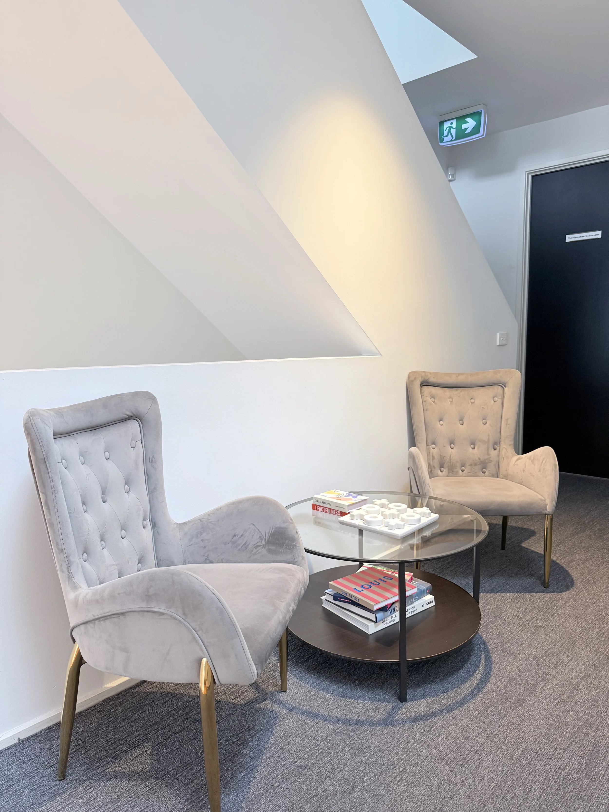 A waiting area with two beige upholstered chairs with button-tufted backs and gold-colored legs, positioned around a round glass coffee table. The table holds a stack of books, a game piece set, and some magazines. The wall behind has a corner with a white sloped ceiling, and an emergency exit sign with a running figure and arrow pointing right is mounted on the wall near a black door. There is a small beige armchair partially visible in the lower left corner.