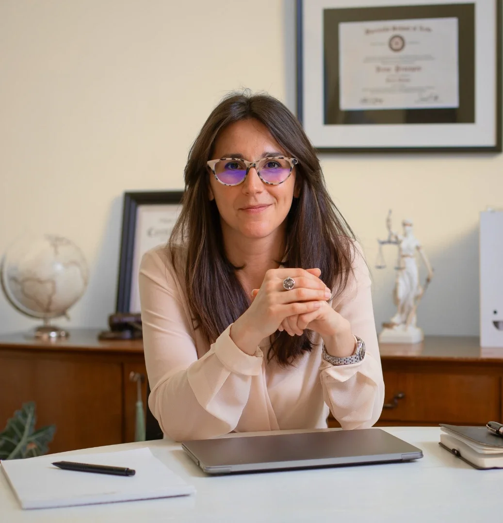 A middle-aged executive woman with brown hair  sits with her elbows on her desk clasping hands while looking directly at the camera with a friendly, but professional smile. She wears glasses and a button up shirt. Her certifications on display