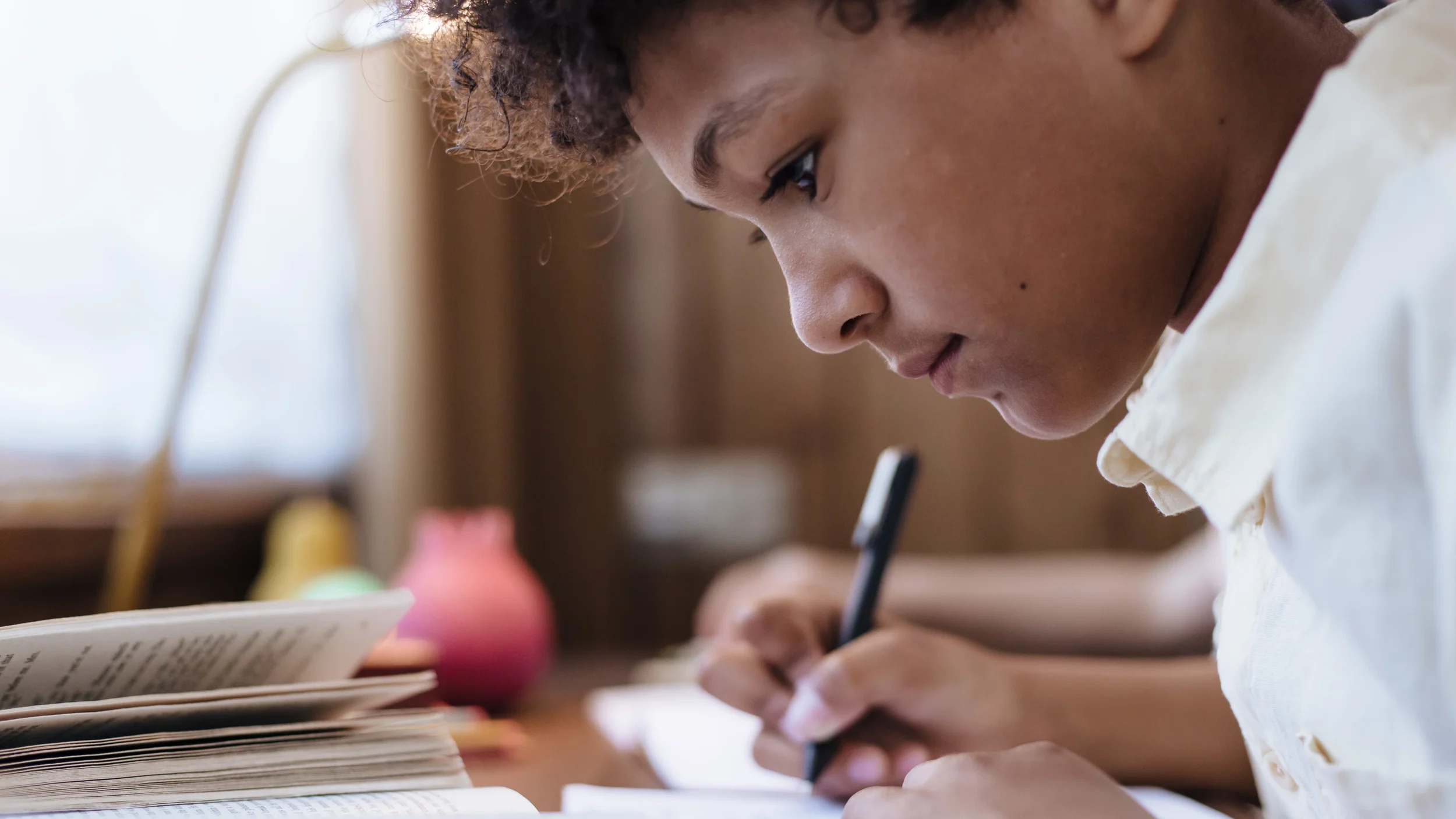 Close-up of a young boy with curly hair concentrating on writing or drawing in a notebook, with books and colorful objects on the table.