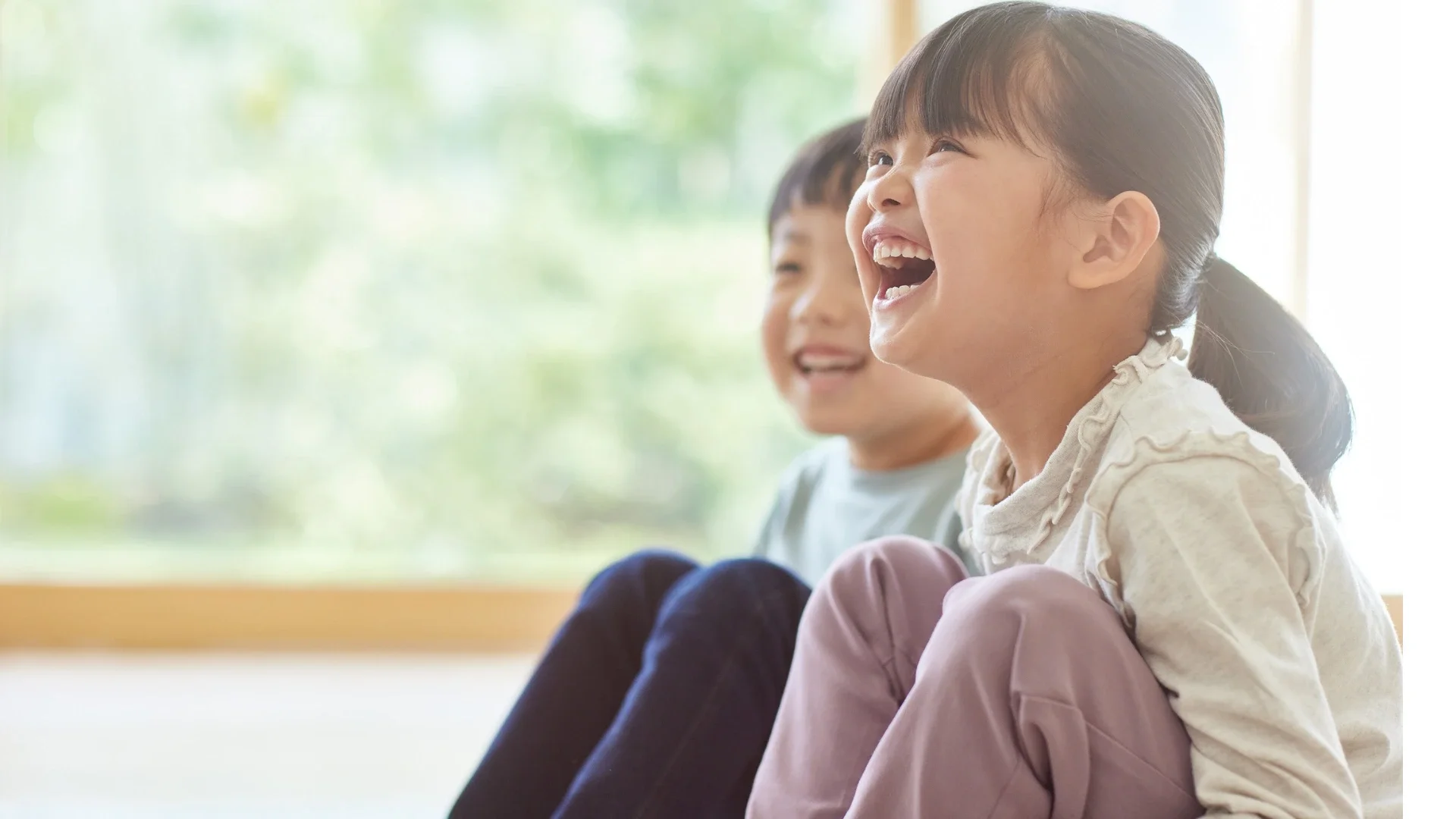 Two young children sitting together, smiling and laughing in a bright room with large windows and greenery outside. Asian decent boy and girl