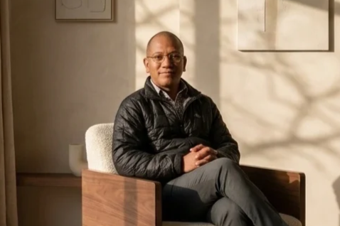 A man sitting on a wooden chair with a cushion, wearing glasses, a black quilted jacket, and gray pants in a warmly lit room.